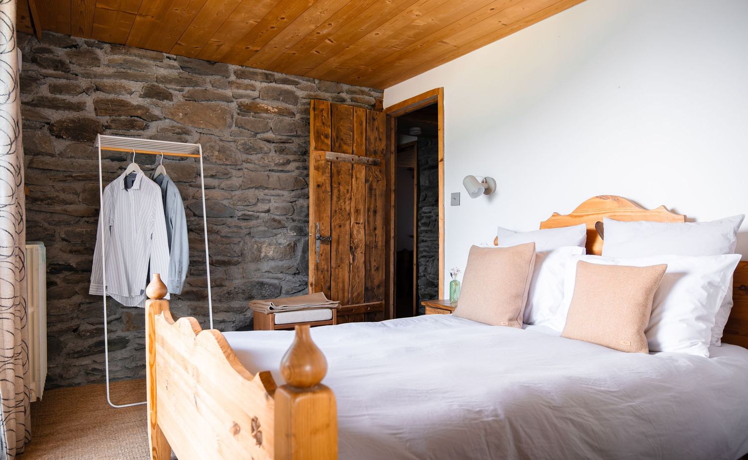 Rustic bedroom with stone wall, wooden ceiling, pine bed frame, white bedding, pink pillows, and clothes rack with hanging shirts.