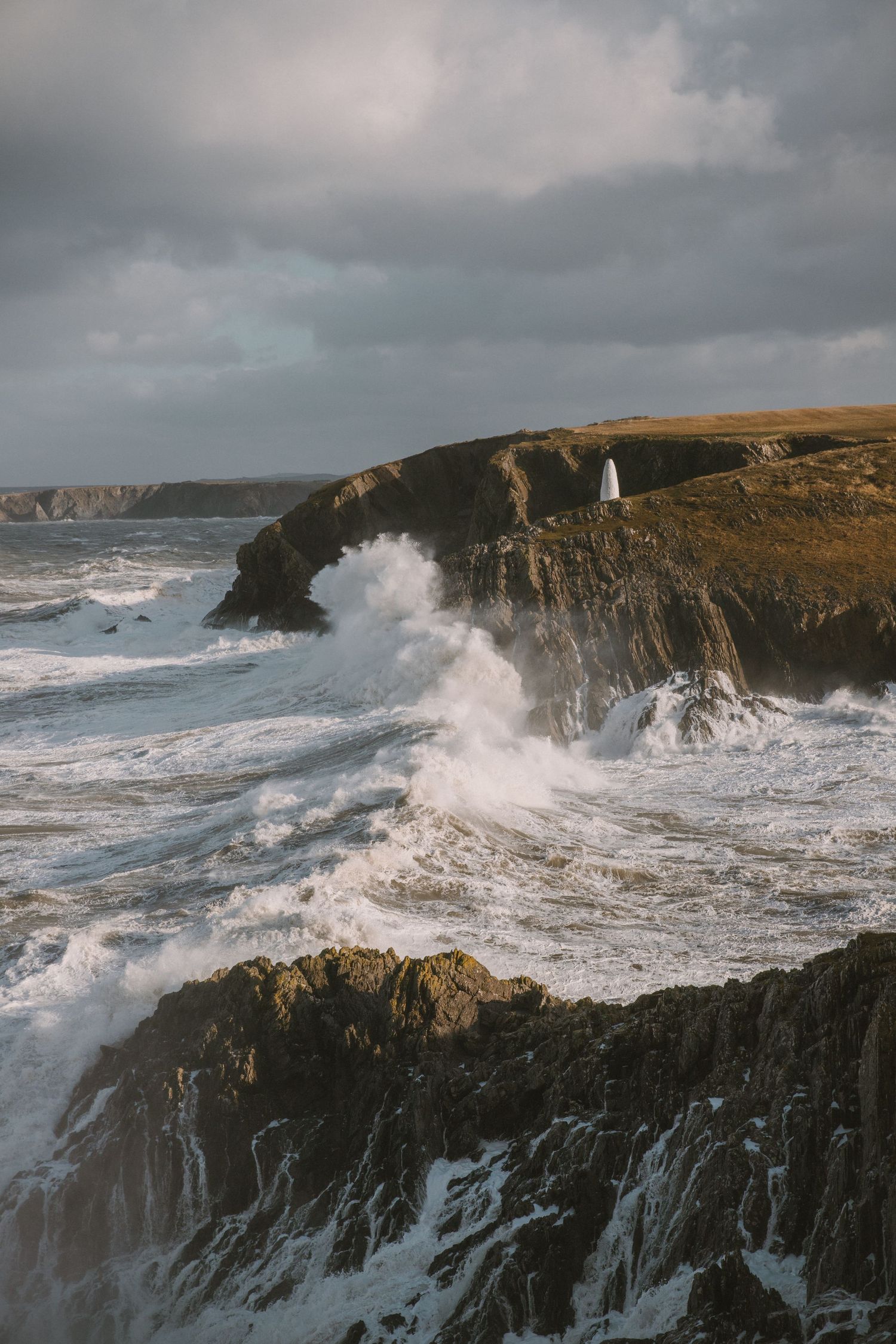 Dramatic ocean waves crash against rugged coastal cliffs under stormy skies at sunset.