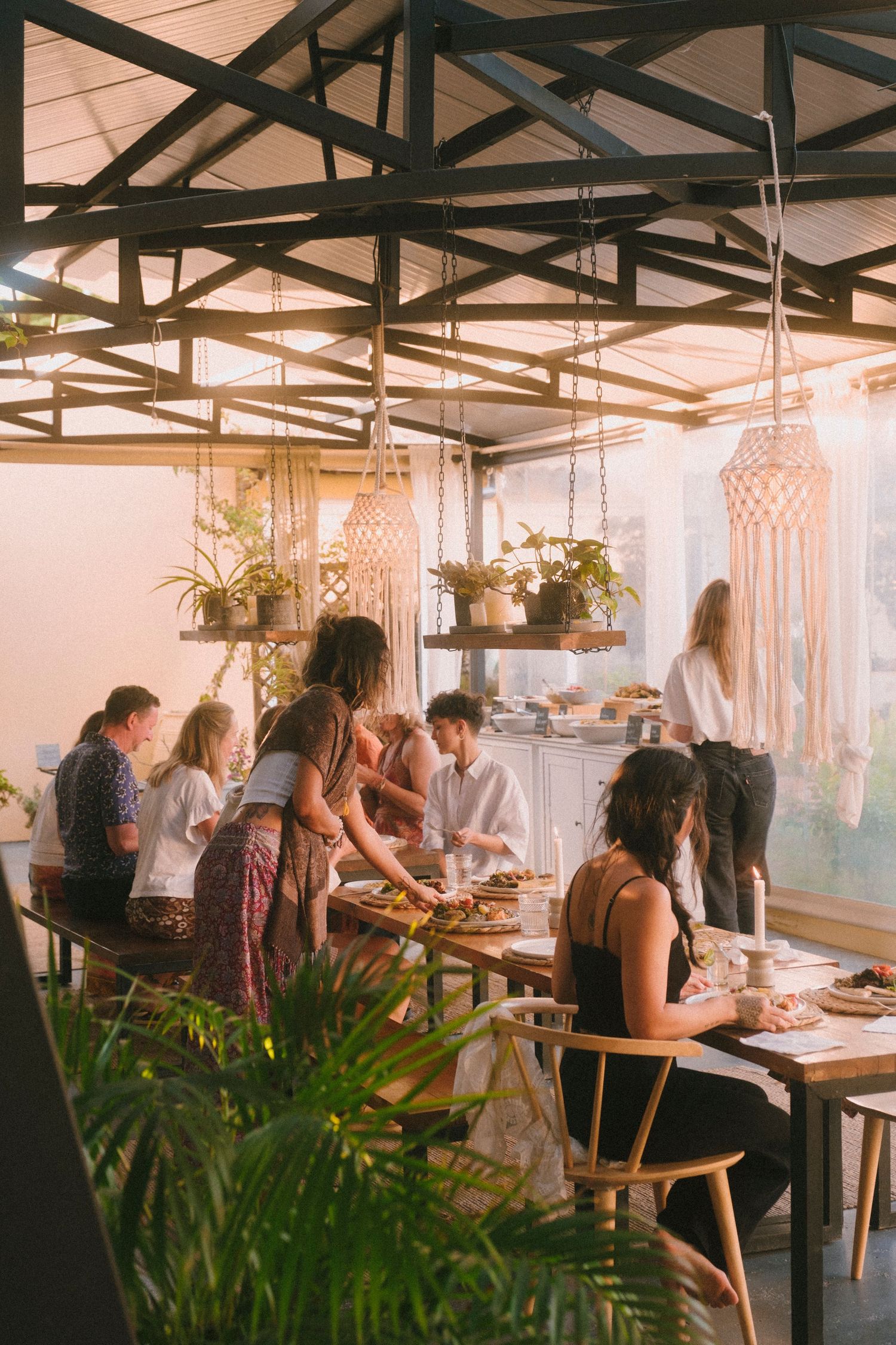 People working and dining in a bright modern restaurant with hanging lamps and tropical plants.