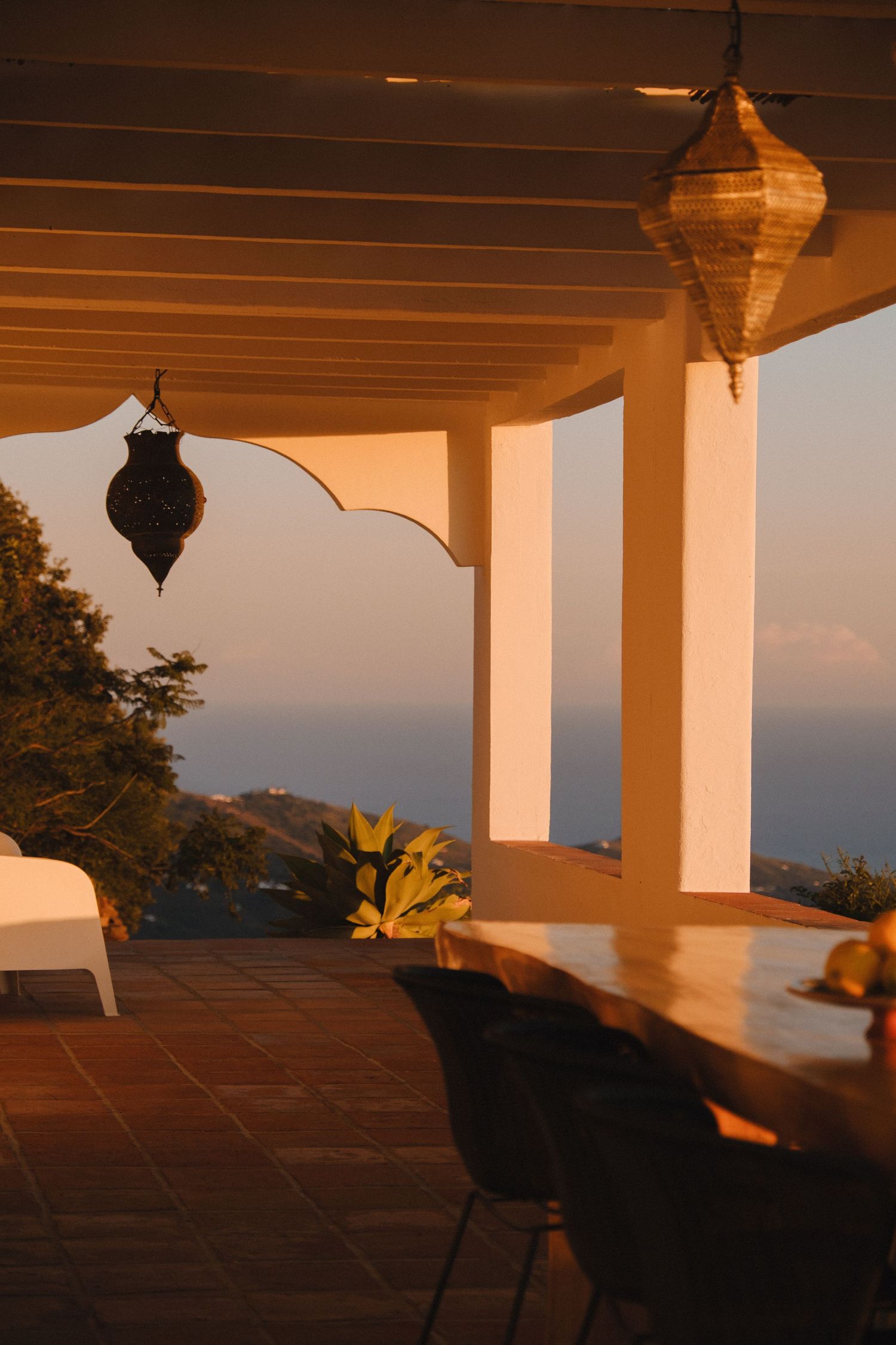Warm sunset light on a Mediterranean terrace with hanging lanterns, white columns, and panoramic sea views.