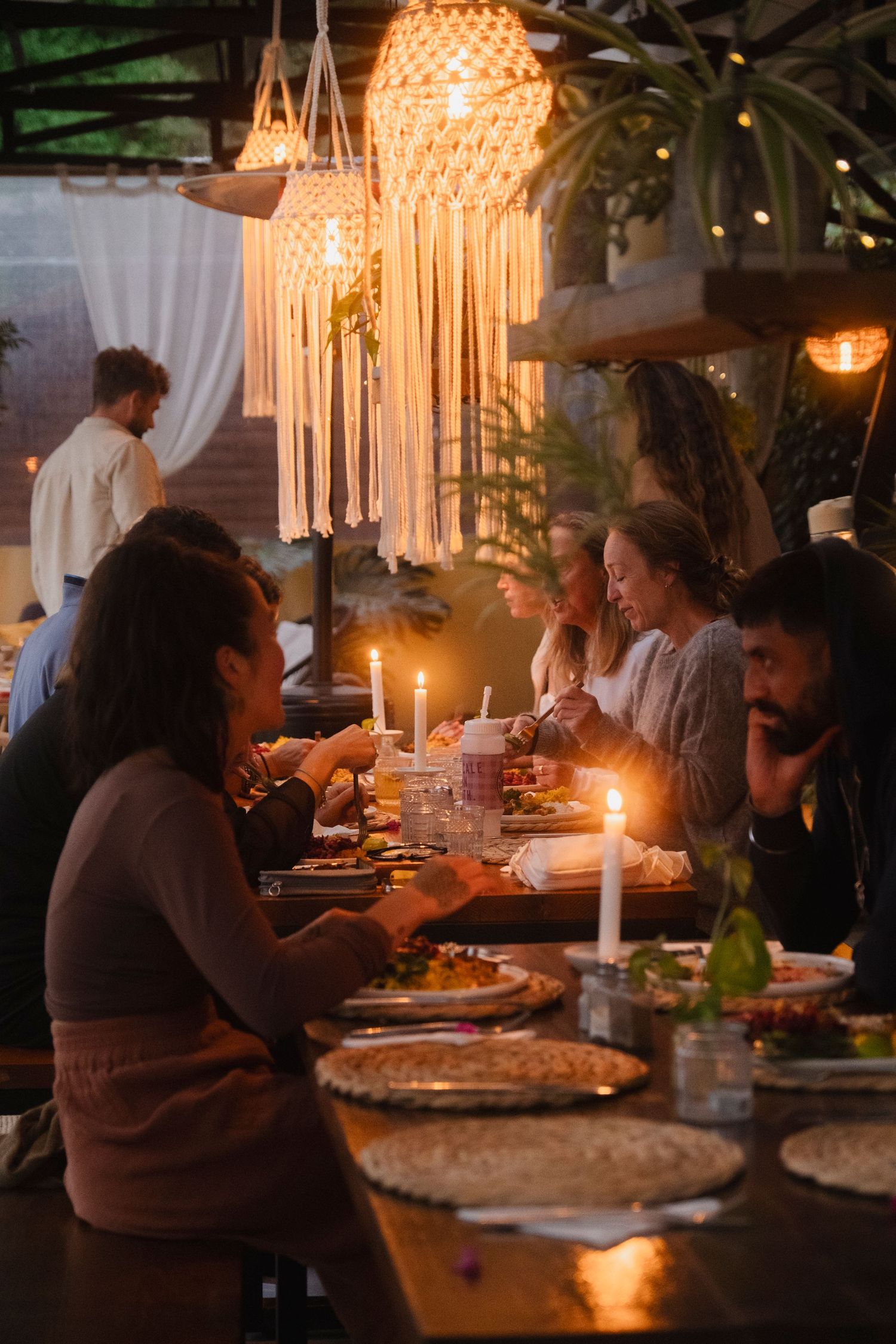 Candlelit dinner gathering with macrame hanging lights creating a warm, intimate atmosphere at a rustic table setting.