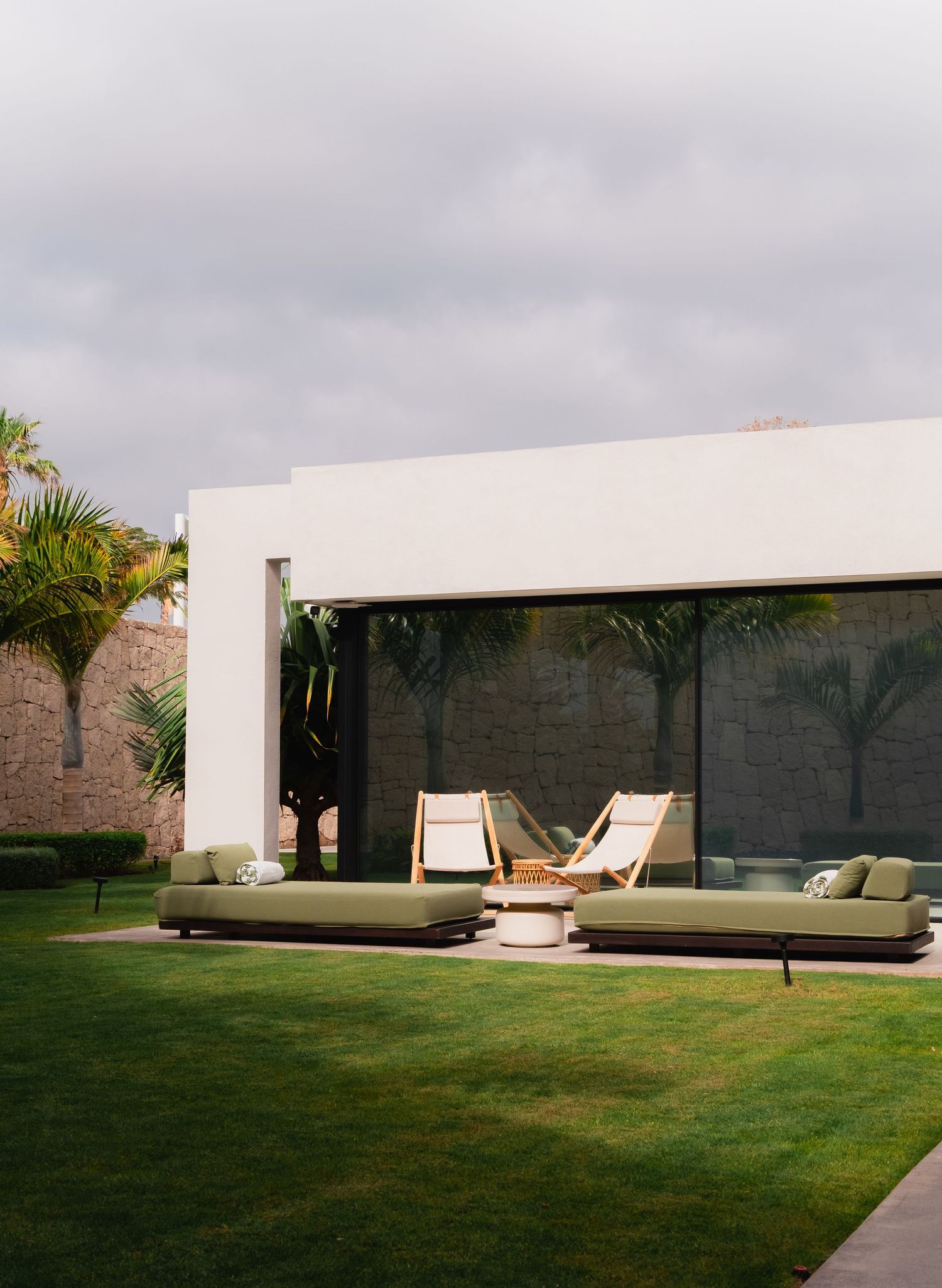 Modern outdoor patio with green sofas, lounge chairs, and lush lawn beside a white minimalist building with palm trees.
