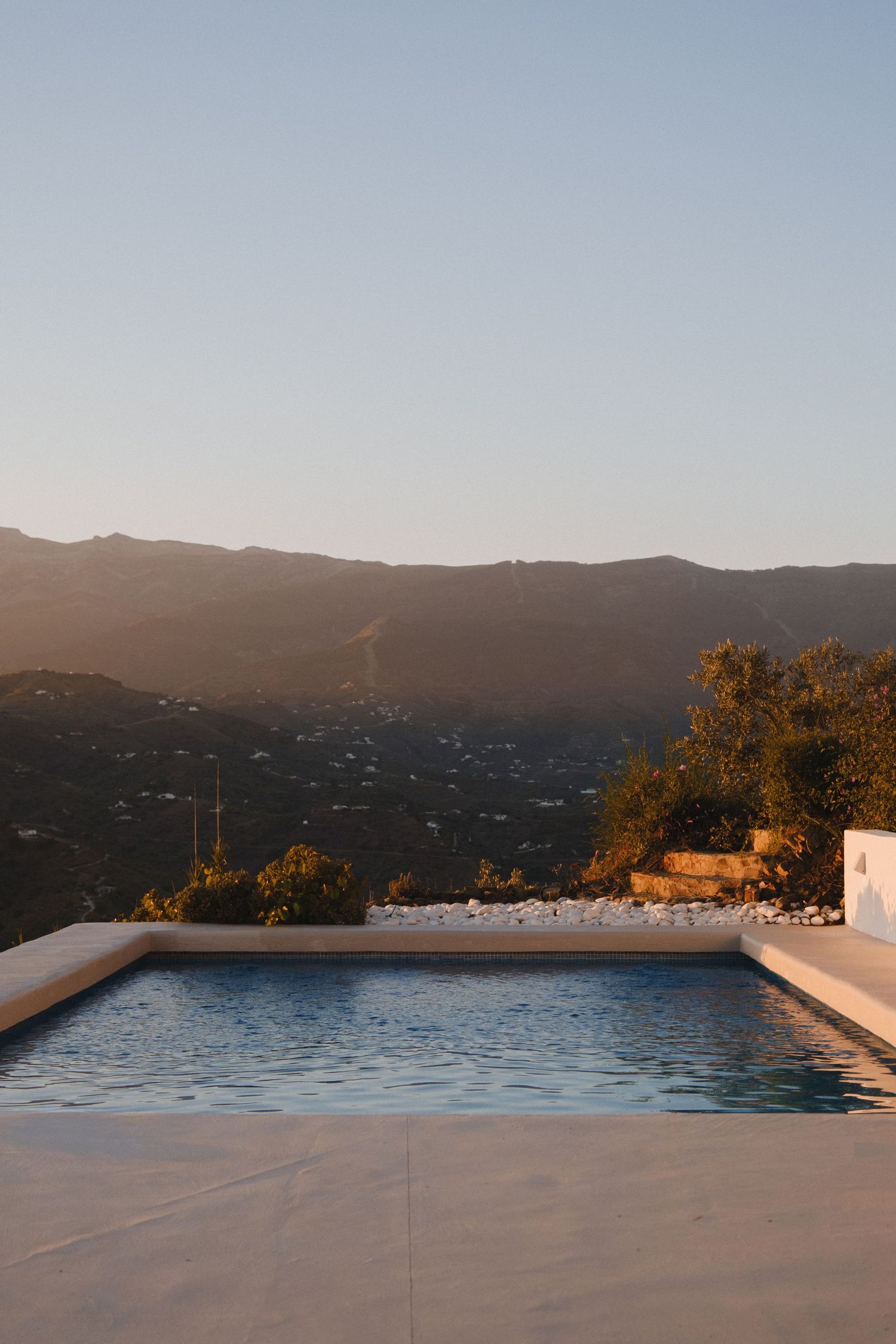 Infinity pool with stunning mountain valley views at golden hour sunset, white stone terrace, Mediterranean landscape.