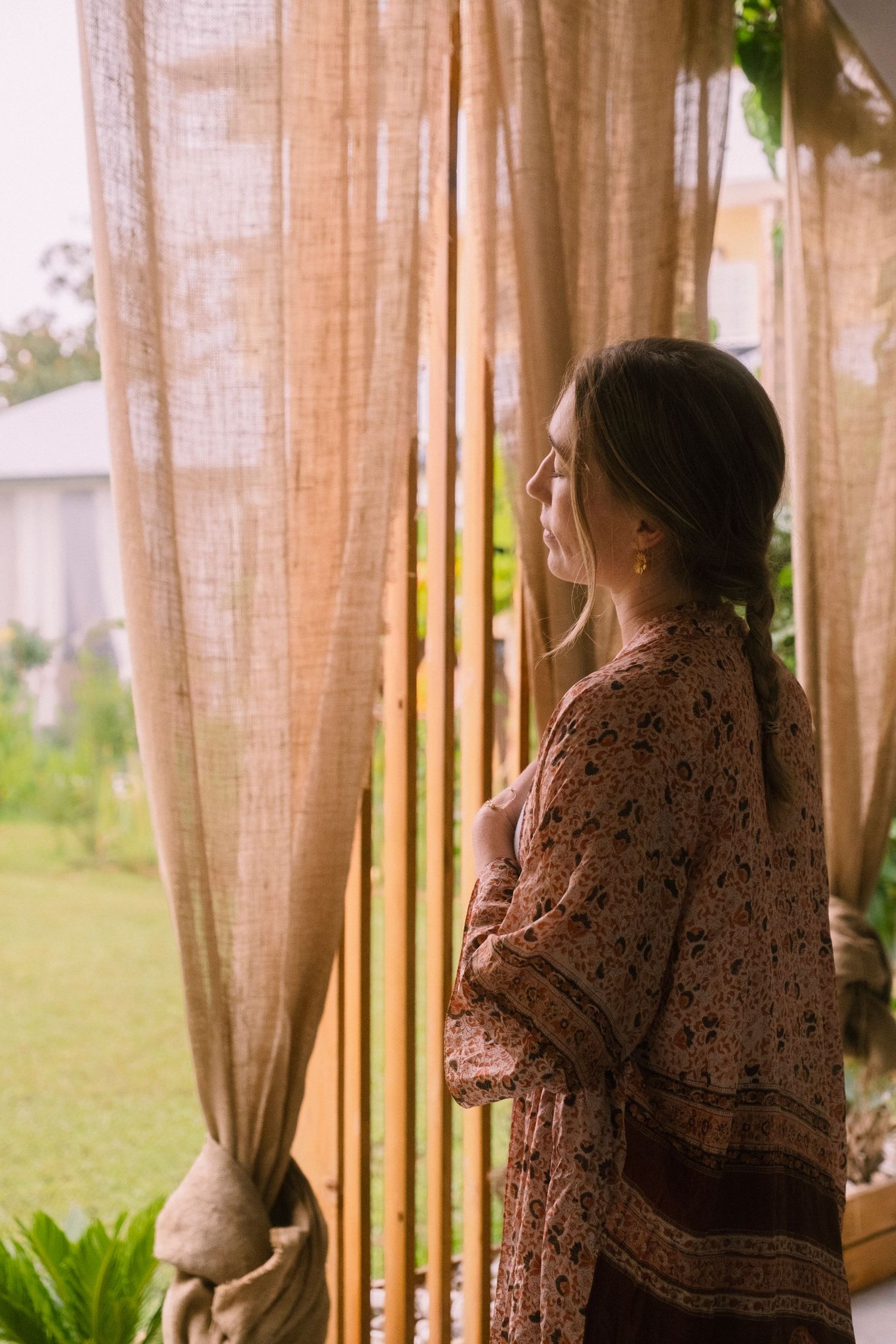A person in a brown patterned dress stands by sheer curtains looking out a window in a contemplative pose.