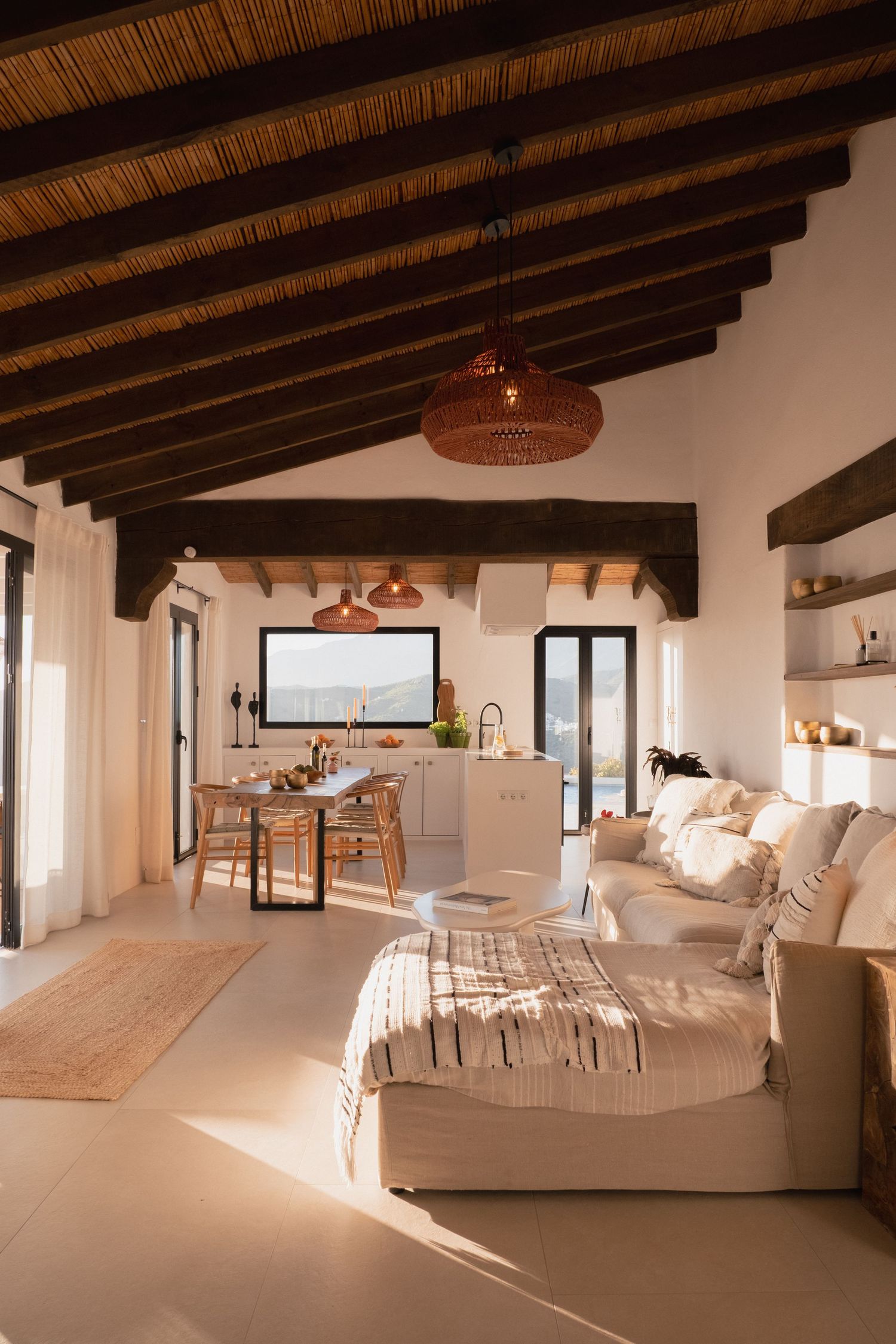 Rustic open-plan studio with exposed wood beam ceiling, white sofa, wicker pendant lights, and a dining area with natural textures.