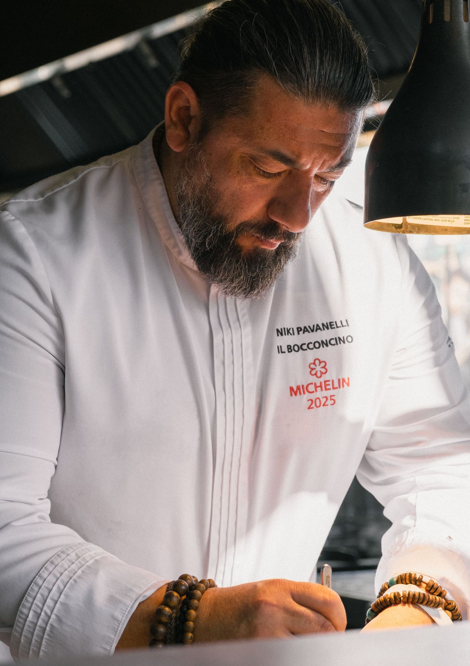 Chef Niki Pavanelli at Il Bocconcino wearing a Michelin 2025 white chef coat, writing at a restaurant table.