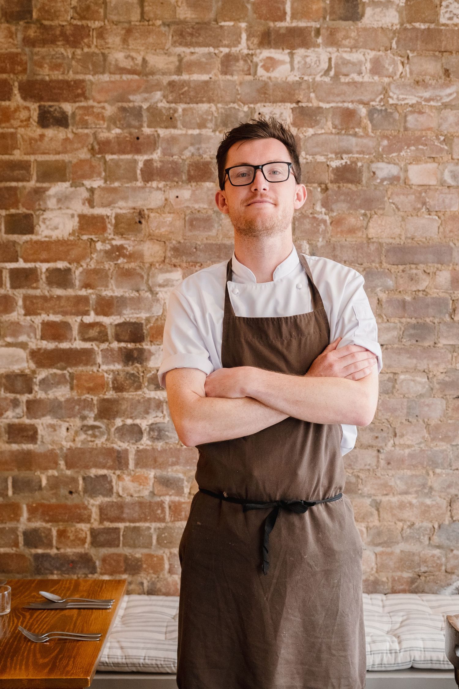 Chef in brown apron standing against exposed brick wall.