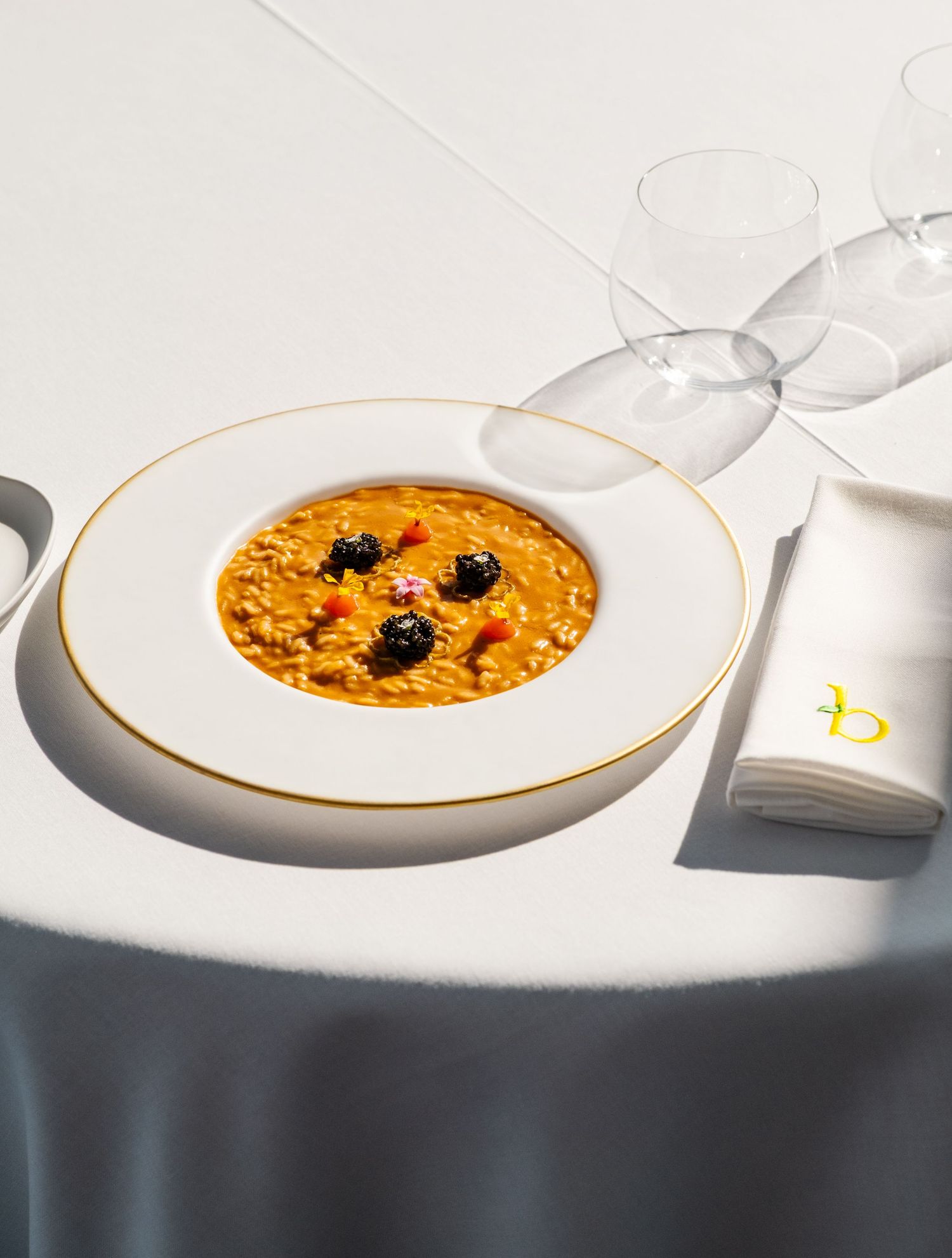 Elegant white and gold rimmed bowl of creamy orange risotto topped with black olives and pink flower garnish.