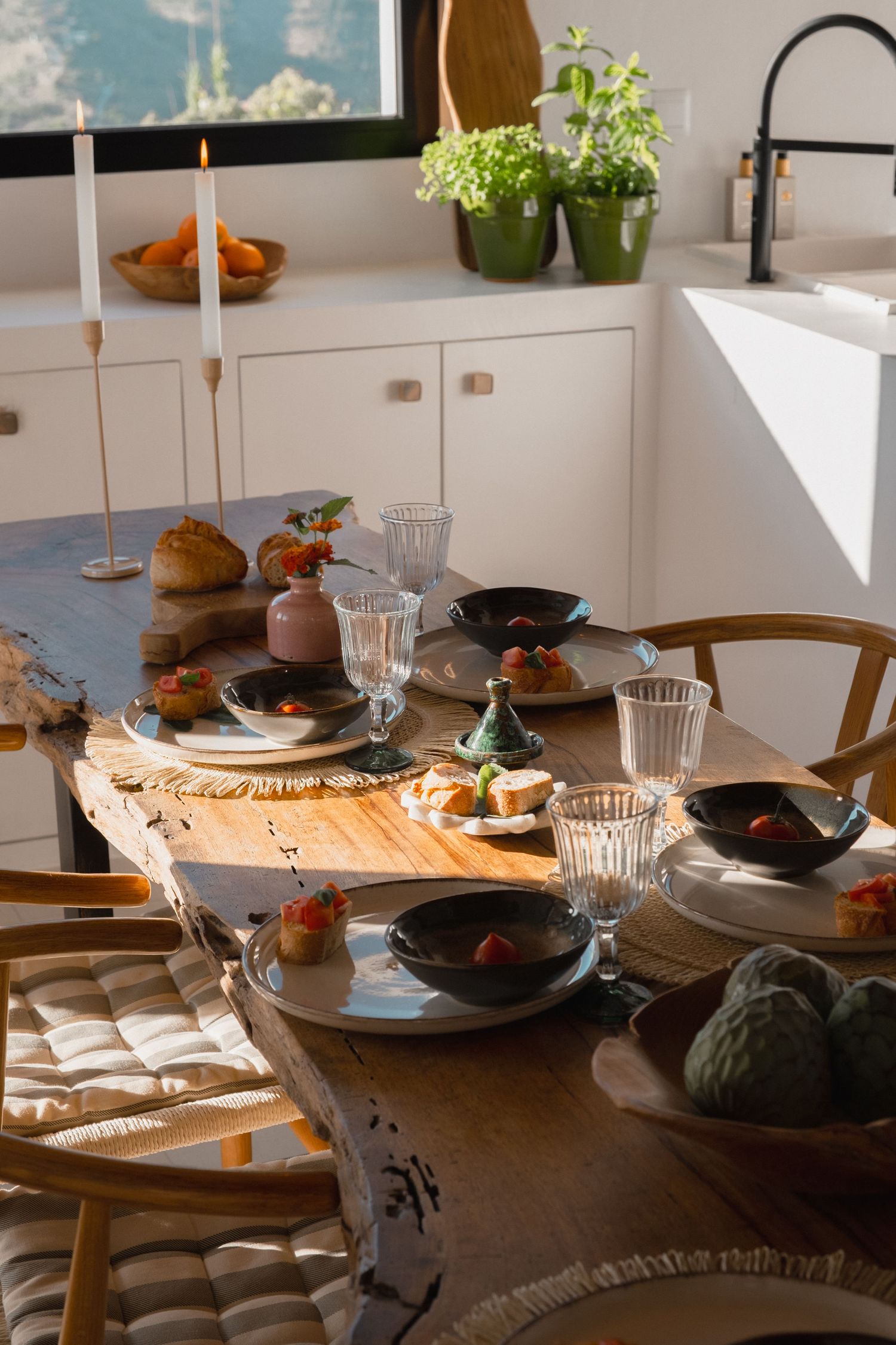 Rustic wooden dining table set with dark ceramic plates, crystal glasses, candles, and fresh food in a bright modern kitchen.