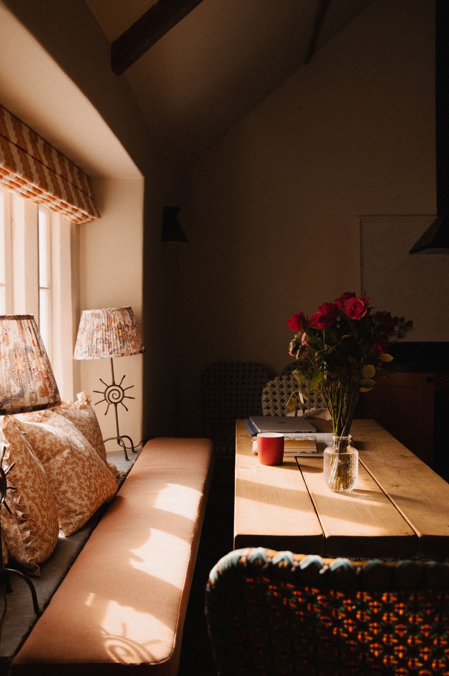 Cozy cottage dining nook with wooden table, red roses in vase, bench seating, and warm sunlight streaming through window.