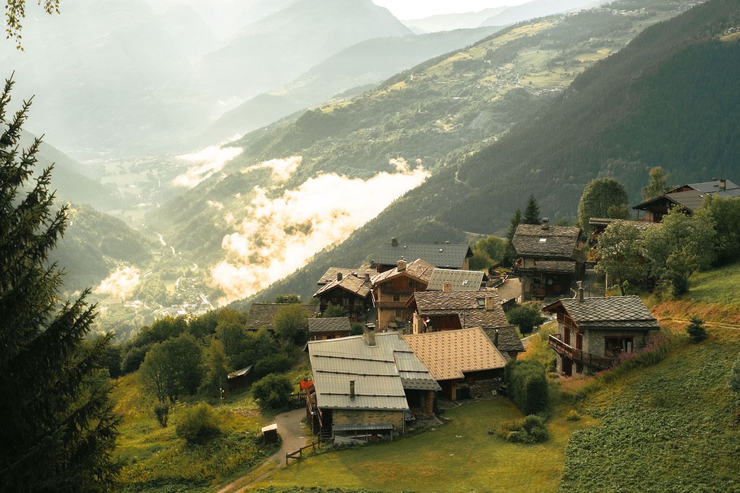 Scenic alpine village with wooden chalets nestled on a lush green hillside surrounded by misty mountain valleys.