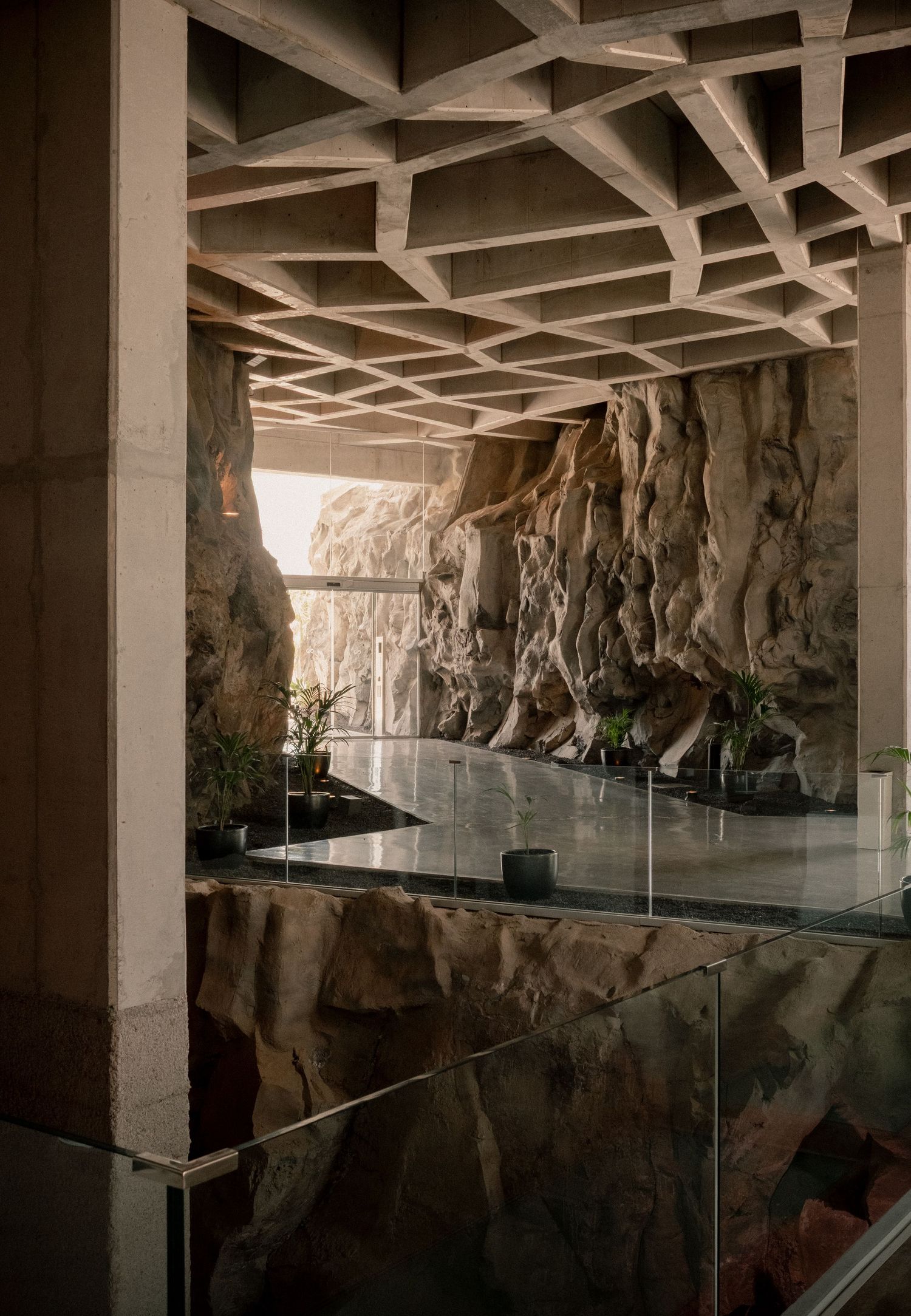 Modern architectural interior with exposed concrete grid ceiling, glass railing, and dramatic natural rock wall feature.