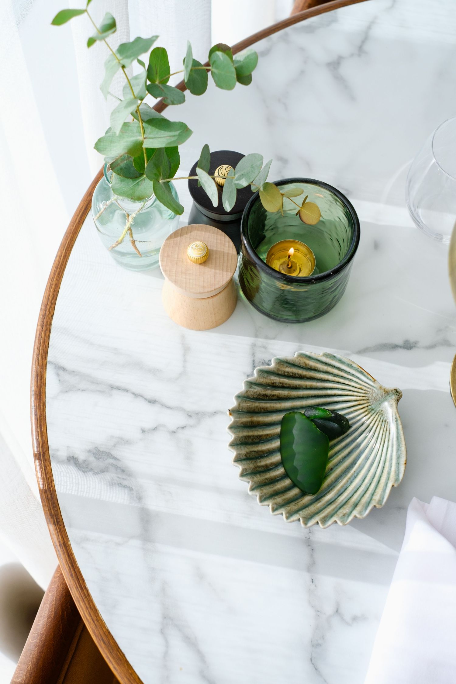 Minimalist marble table setting with eucalyptus, green glass vessels, and seashell decor.