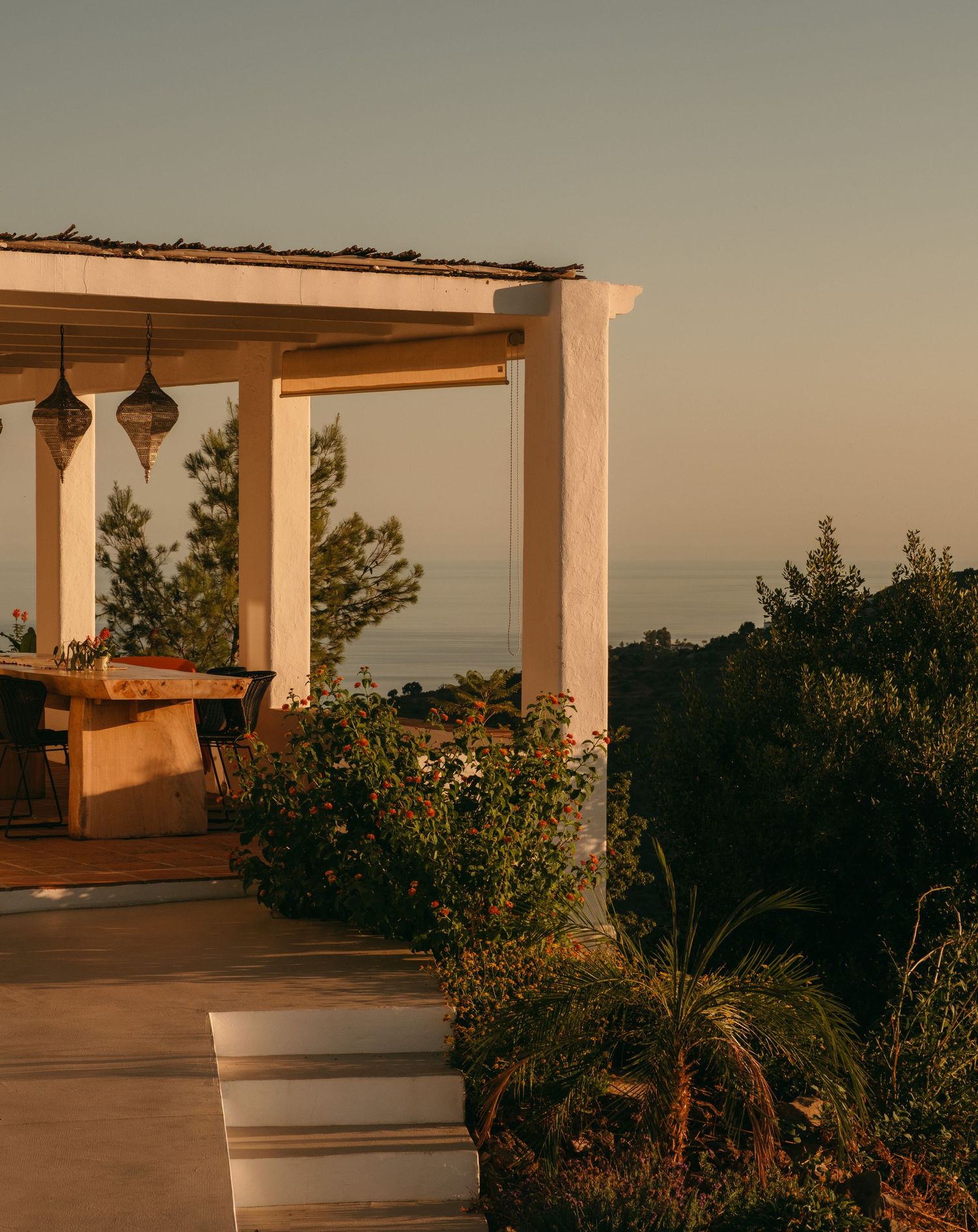 A sunlit Mediterranean villa terrace with white columns, hanging lanterns, and panoramic sea views at golden hour.
