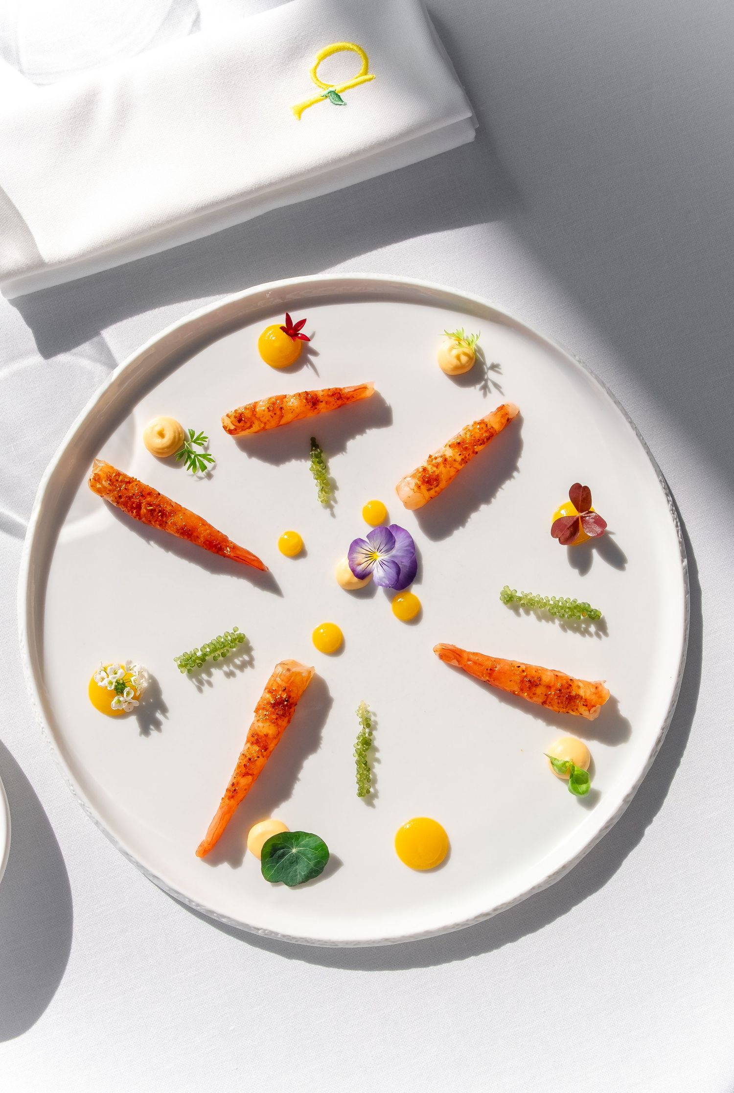 Elegant fine dining plate with roasted baby carrots, edible flowers, microgreens, and yellow sauce dots on white ceramic.