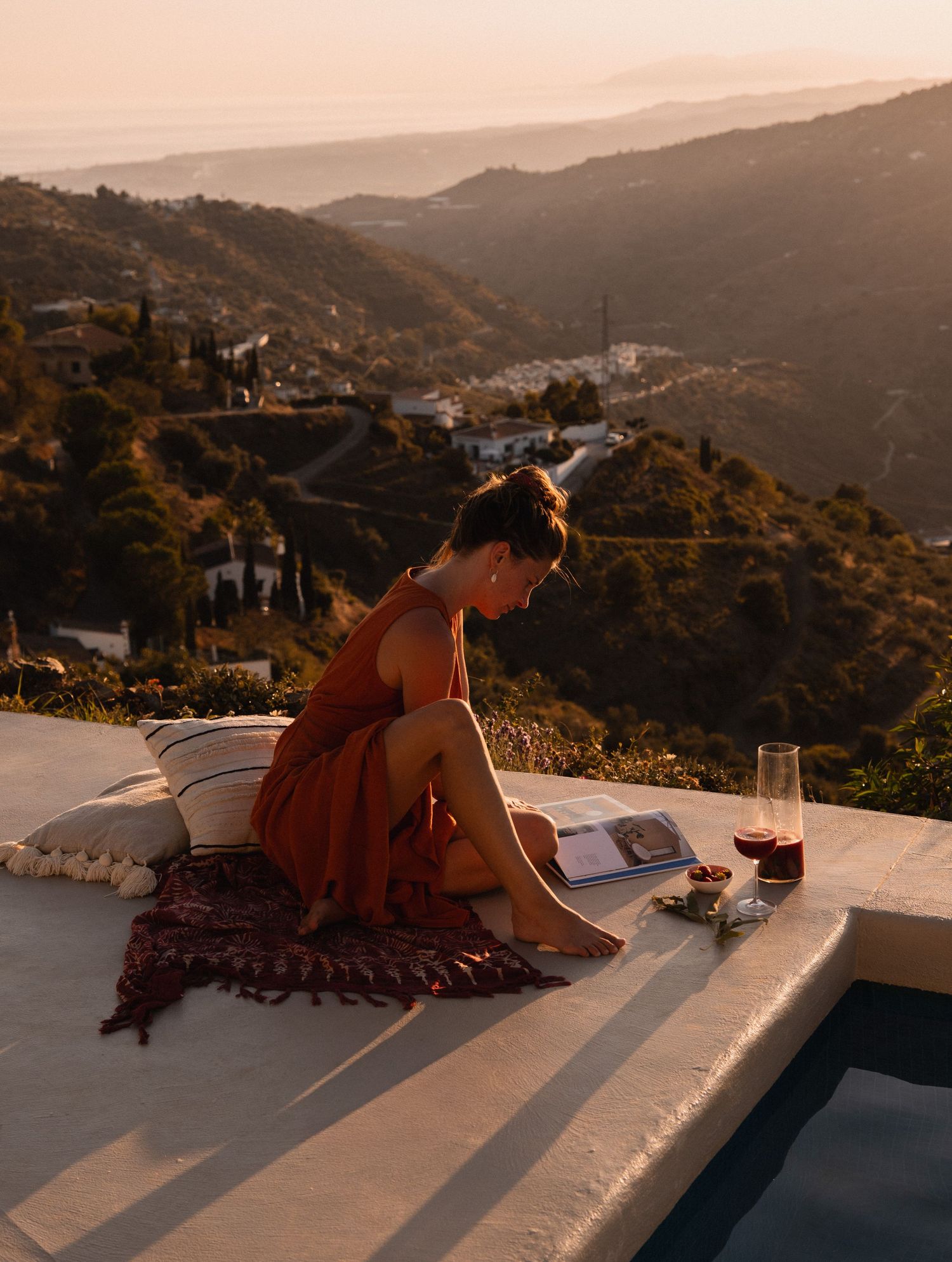 Woman in orange dress reading by a pool at sunset with scenic mountain valley views in the background.