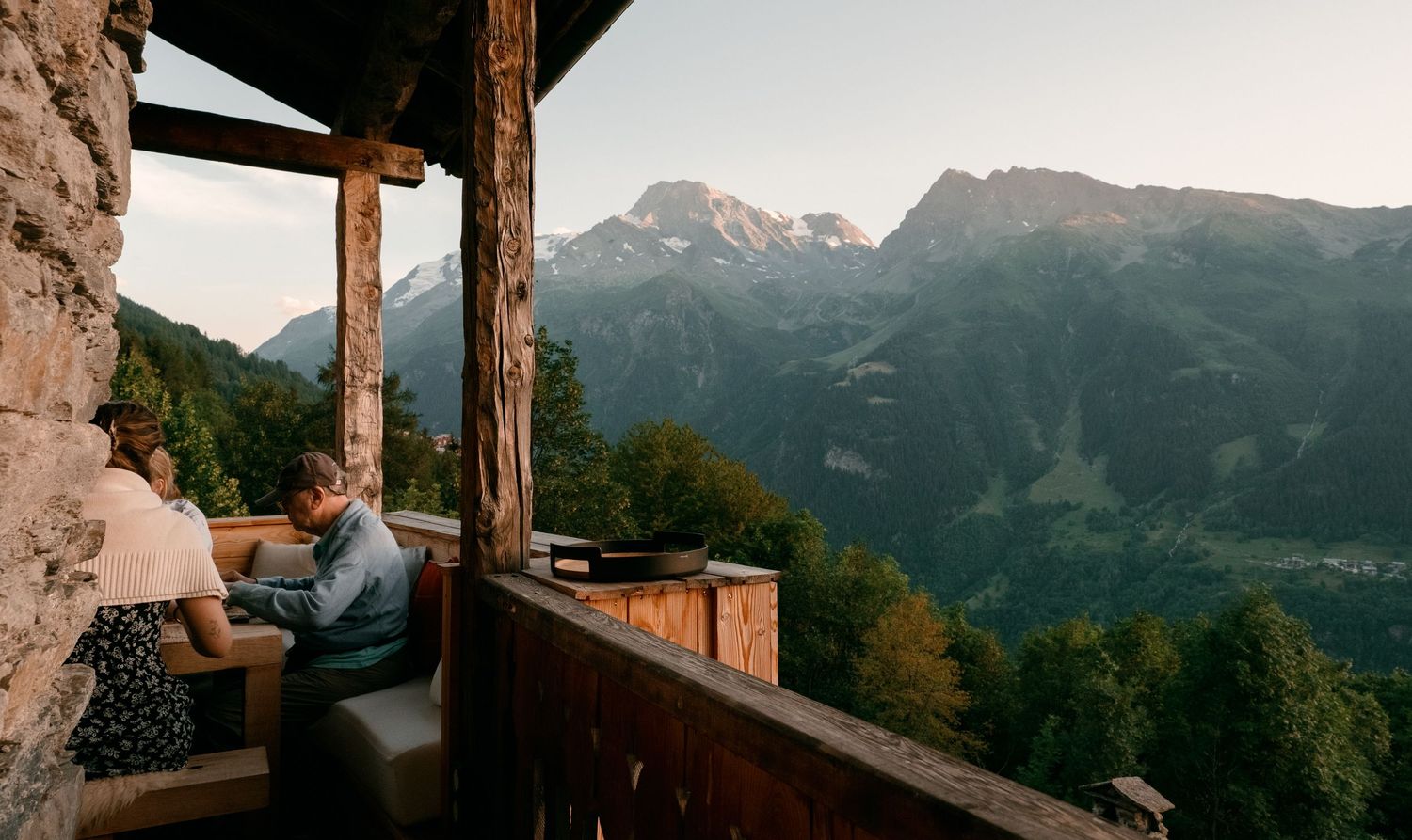 Couple relaxing on a rustic mountain chalet balcony with stunning Alpine peaks and forested valley views at dusk.