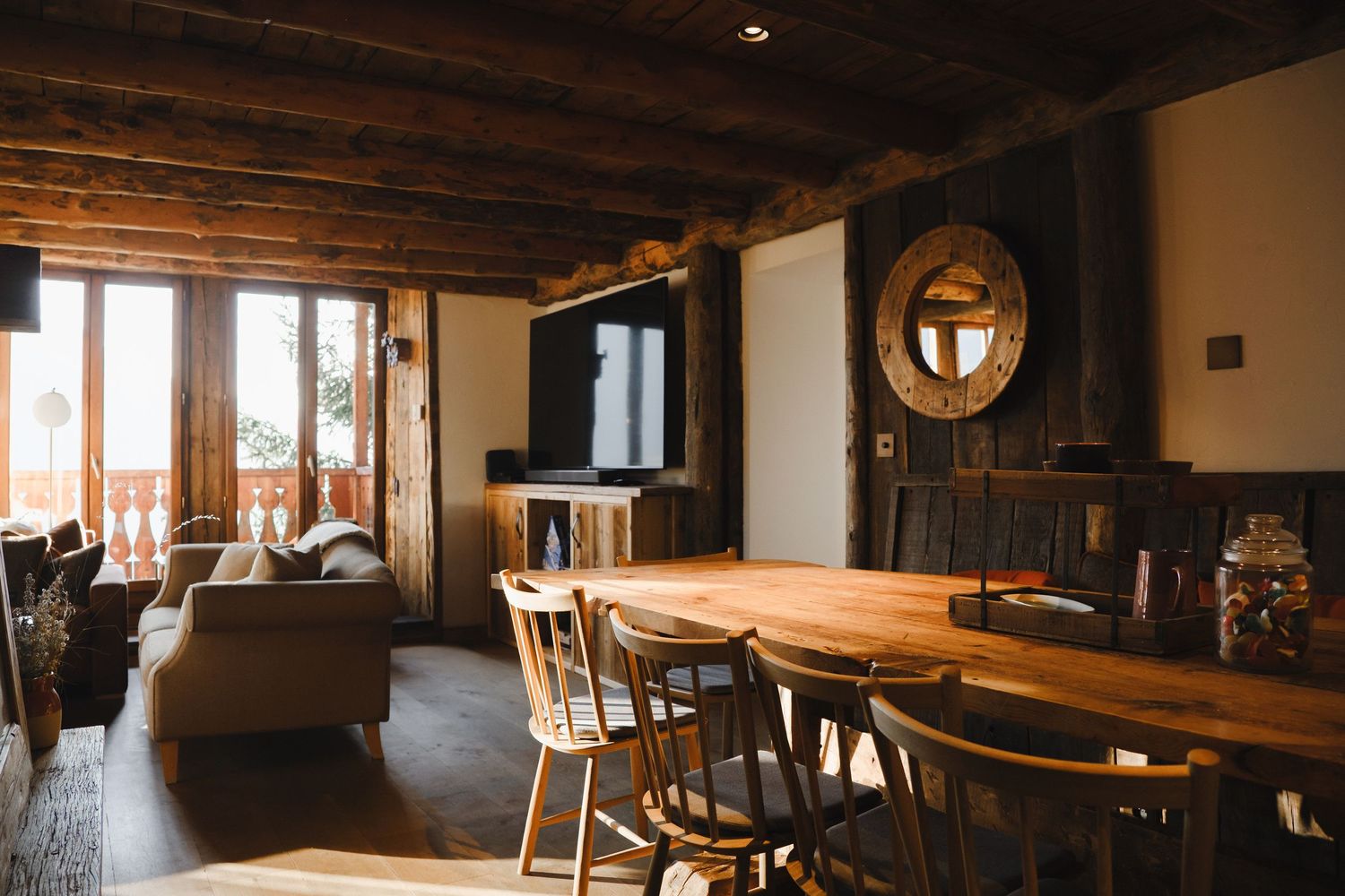 Rustic chalet dining room with wooden table, chairs, exposed beam ceiling, and cozy living area in background.