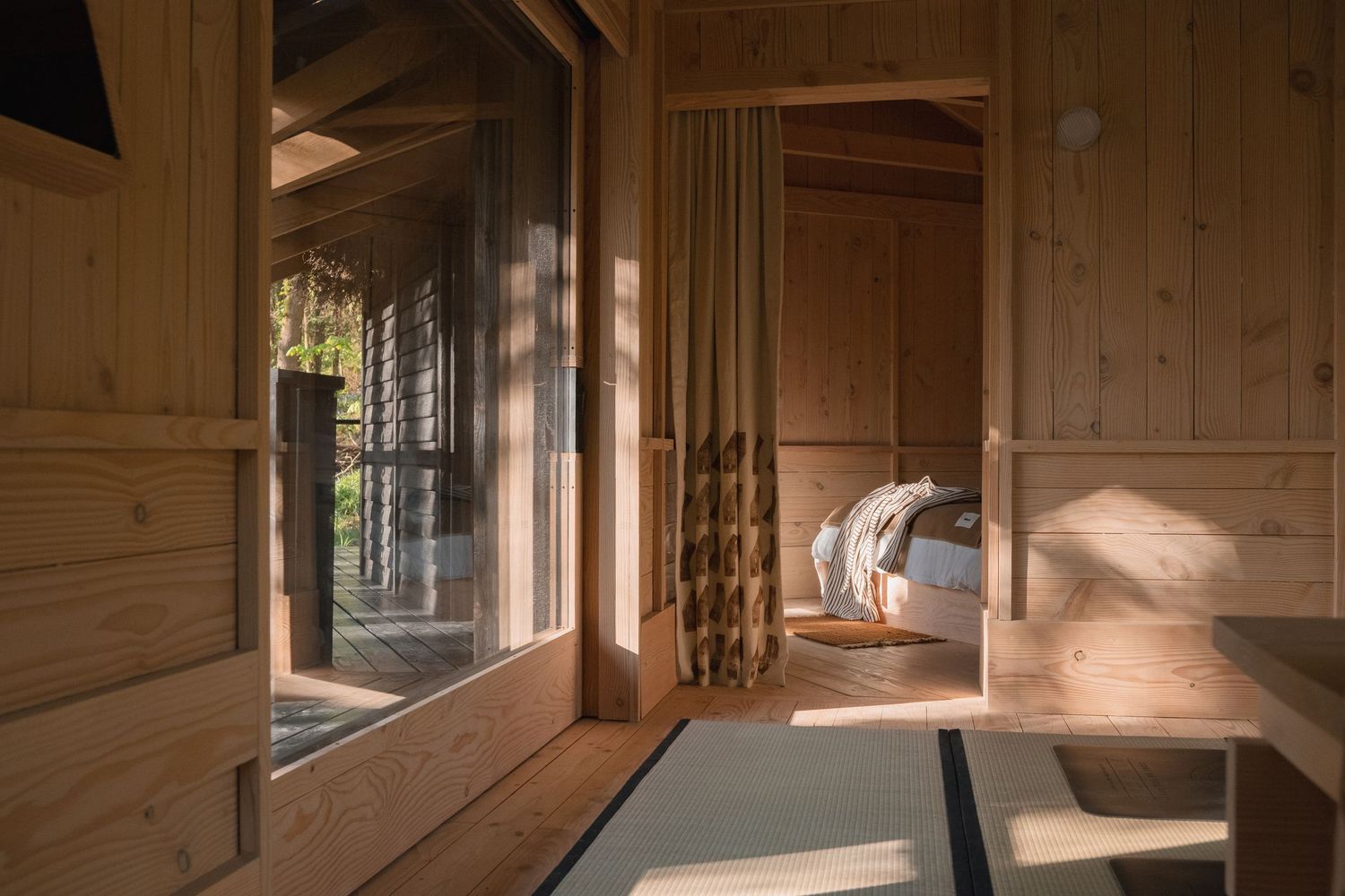 Warm wooden cabin interior with tatami floor, sheer curtains, and sunlit bedroom visible through open doorway.