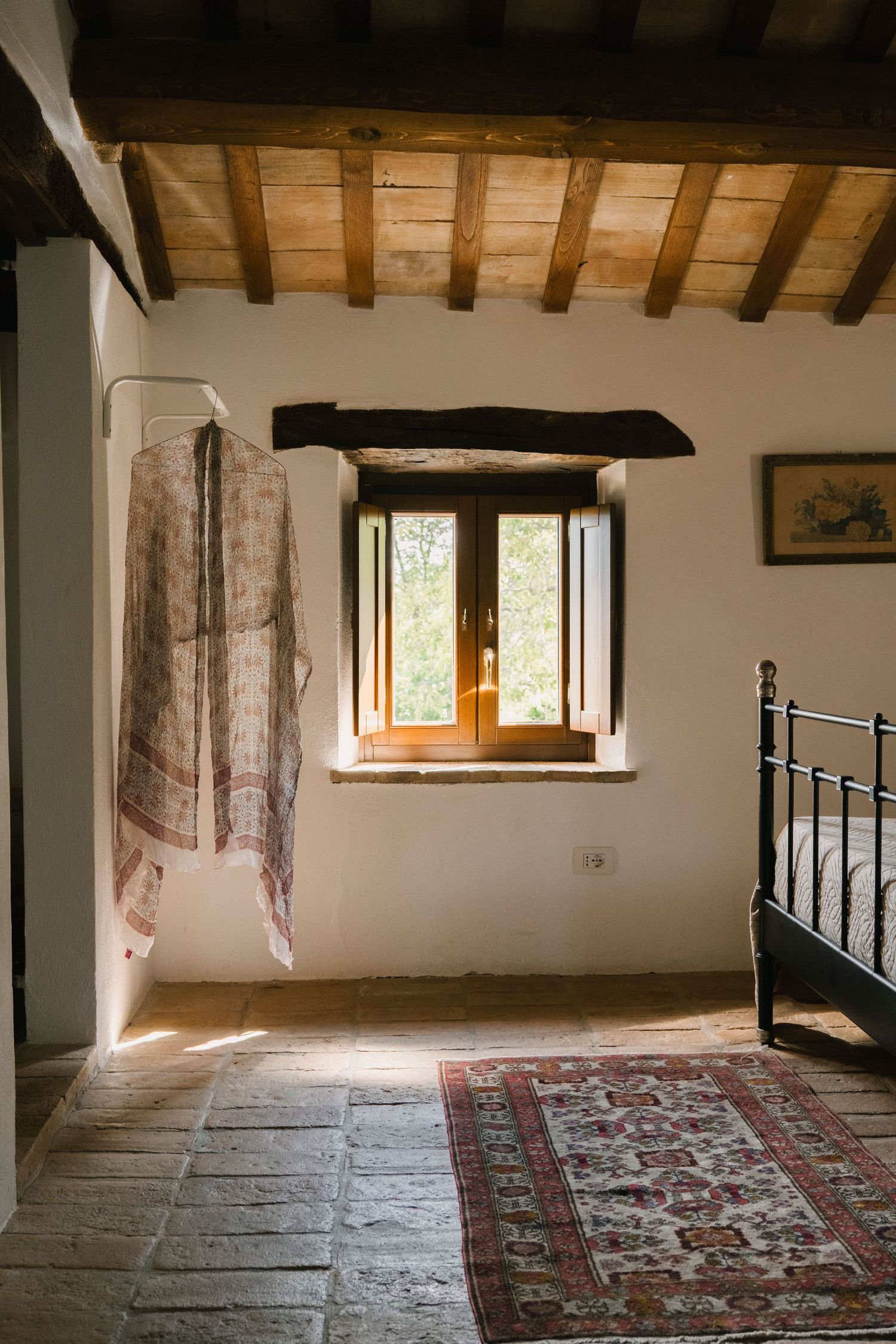 Rustic Tuscan bedroom with exposed wooden beams, stone floor, Persian rug, iron bed, and sunlit window.