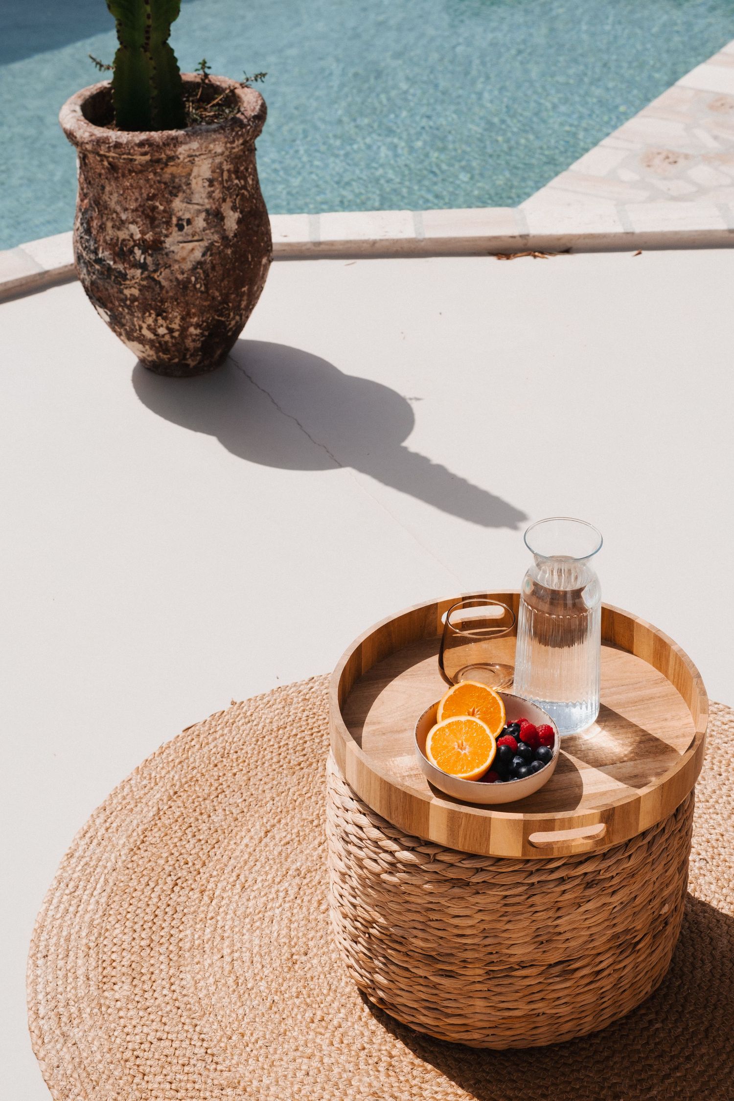Woven rattan tray with oranges and glass of water by poolside on white surface.