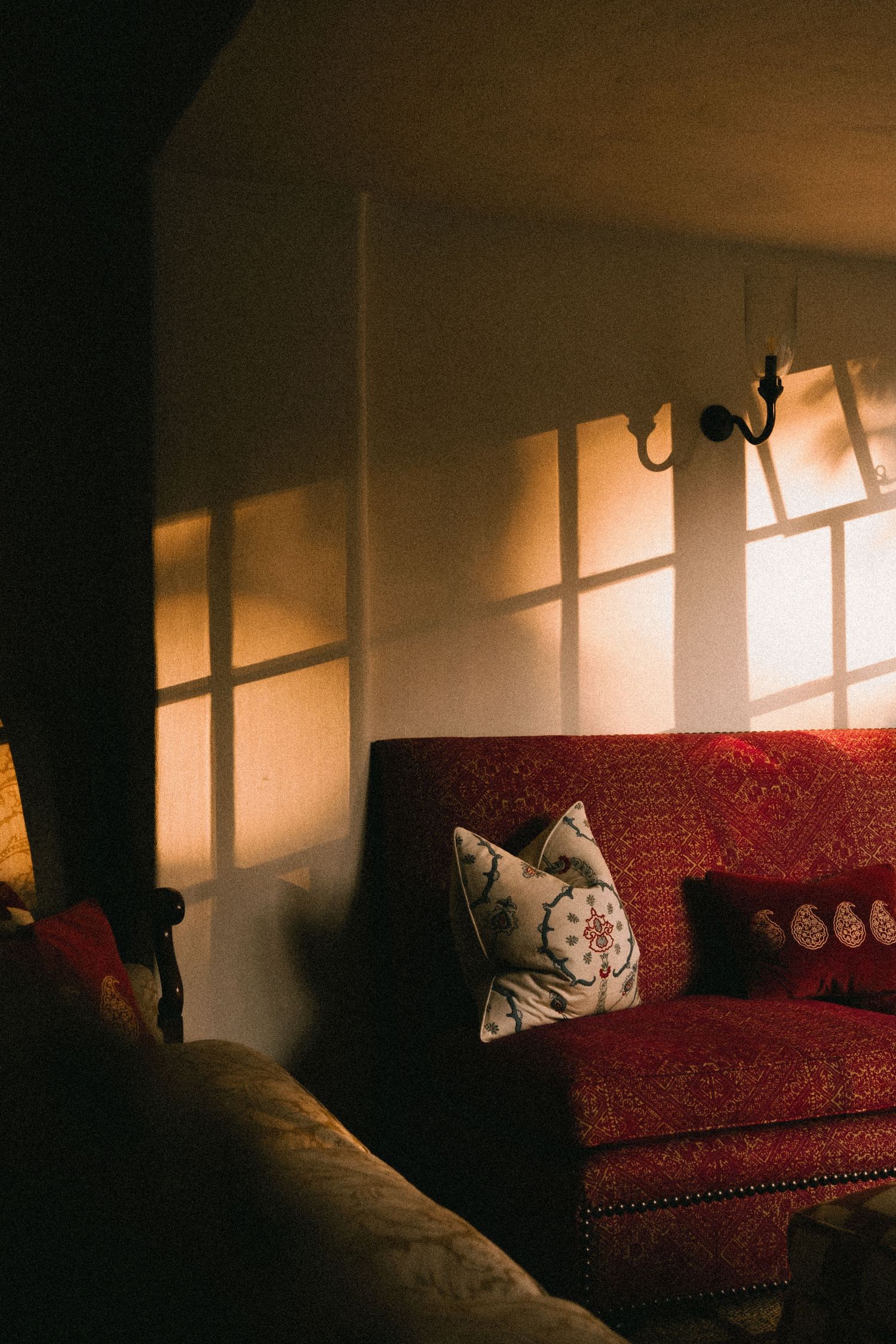 Warm sunlight casts window shadows on a beige wall above a red patterned sofa with decorative pillows in a cozy living room.