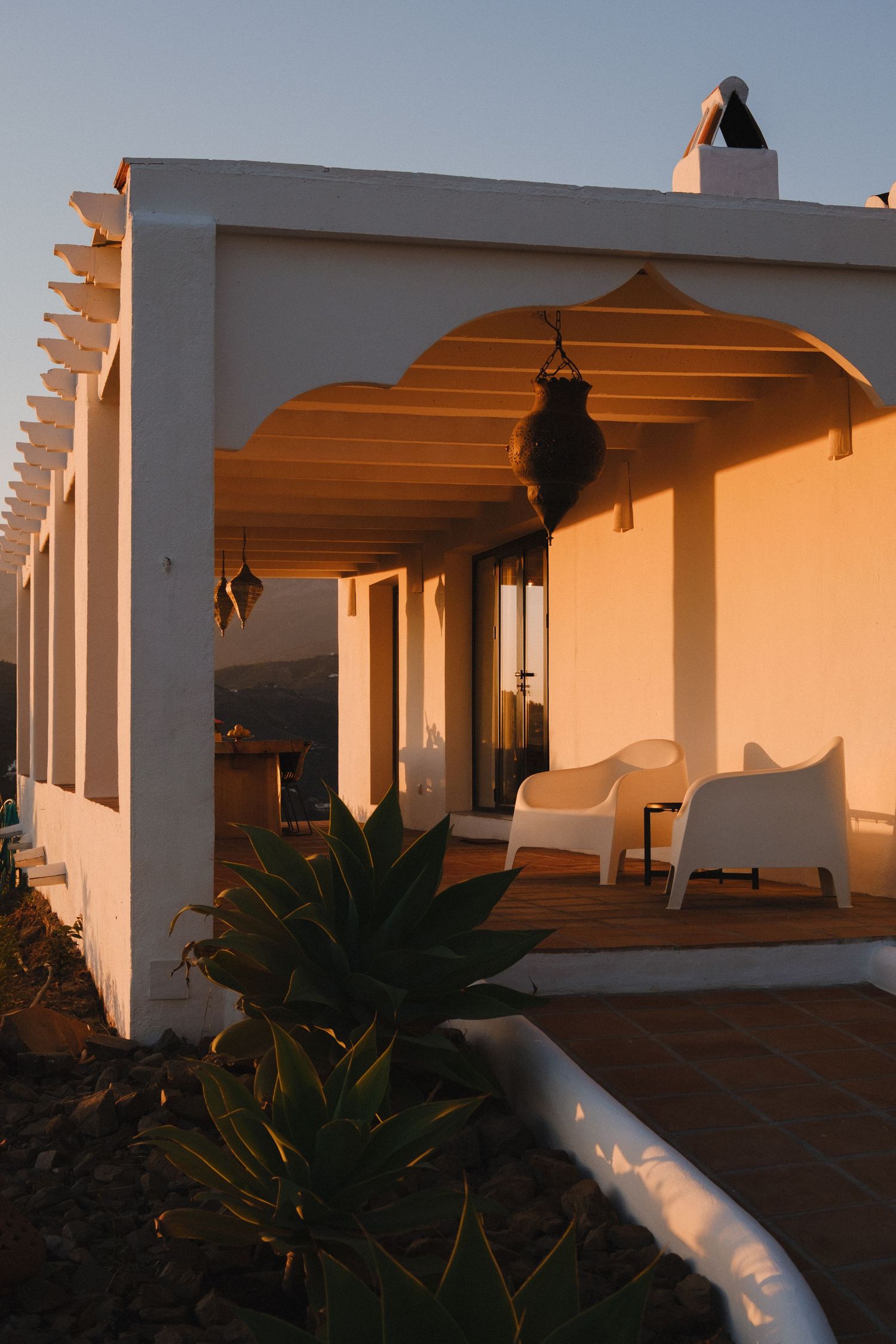 White Mediterranean villa patio with arched pergola bathed in golden sunset light, featuring white chairs and agave plants.