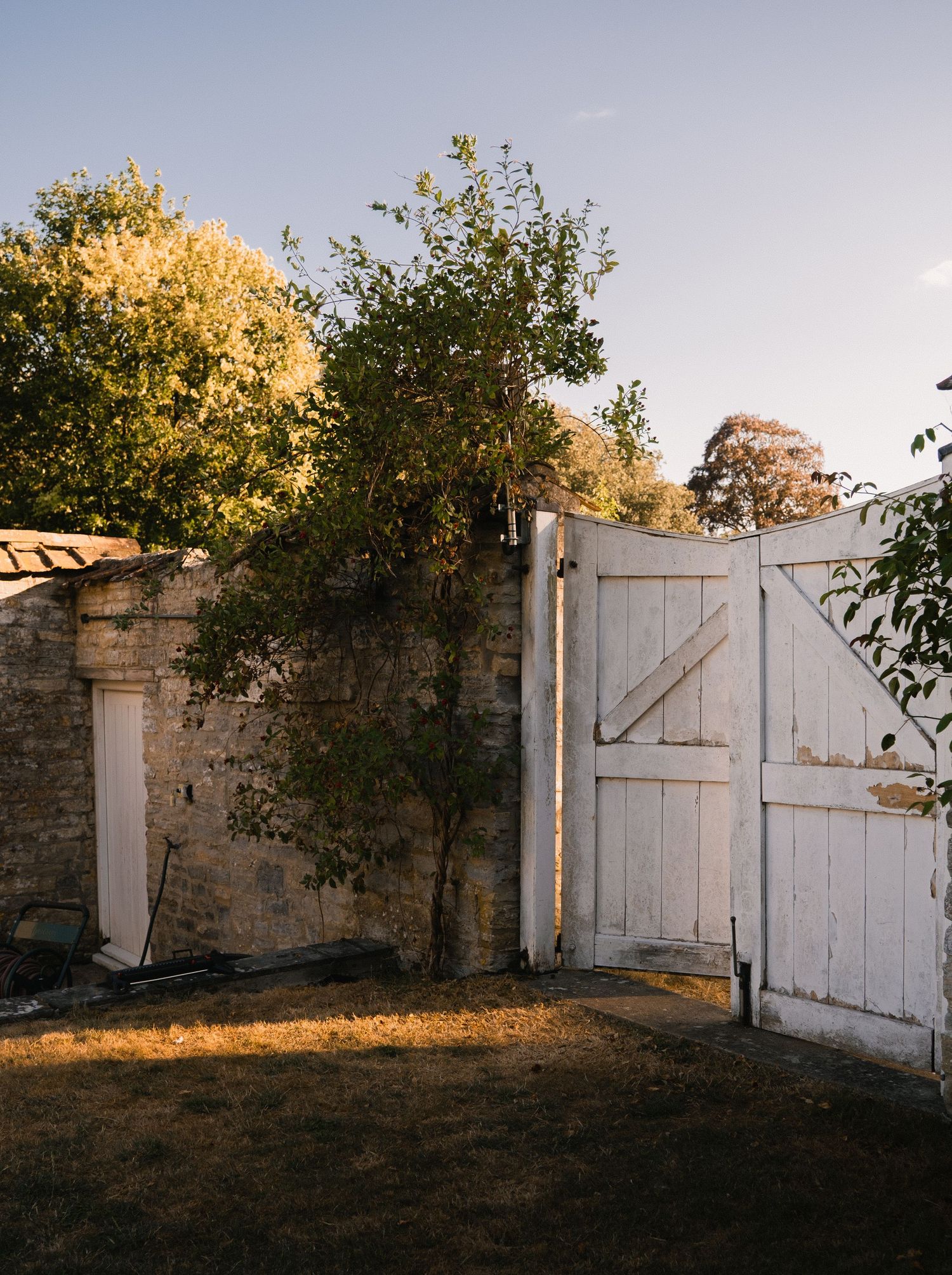 A rustic white wooden gate beside a stone wall with lush green trees in a sunlit garden.