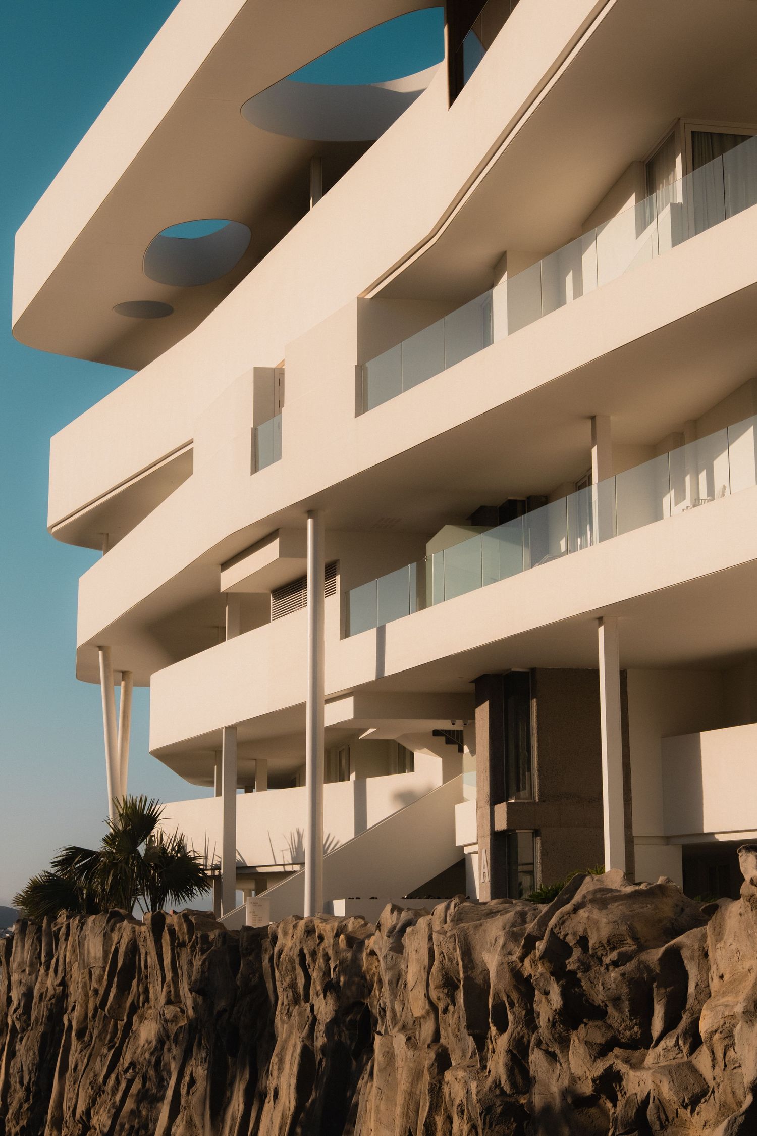 Modern white brutalist building with curved balconies perched on rocky cliffs under a clear blue sky.