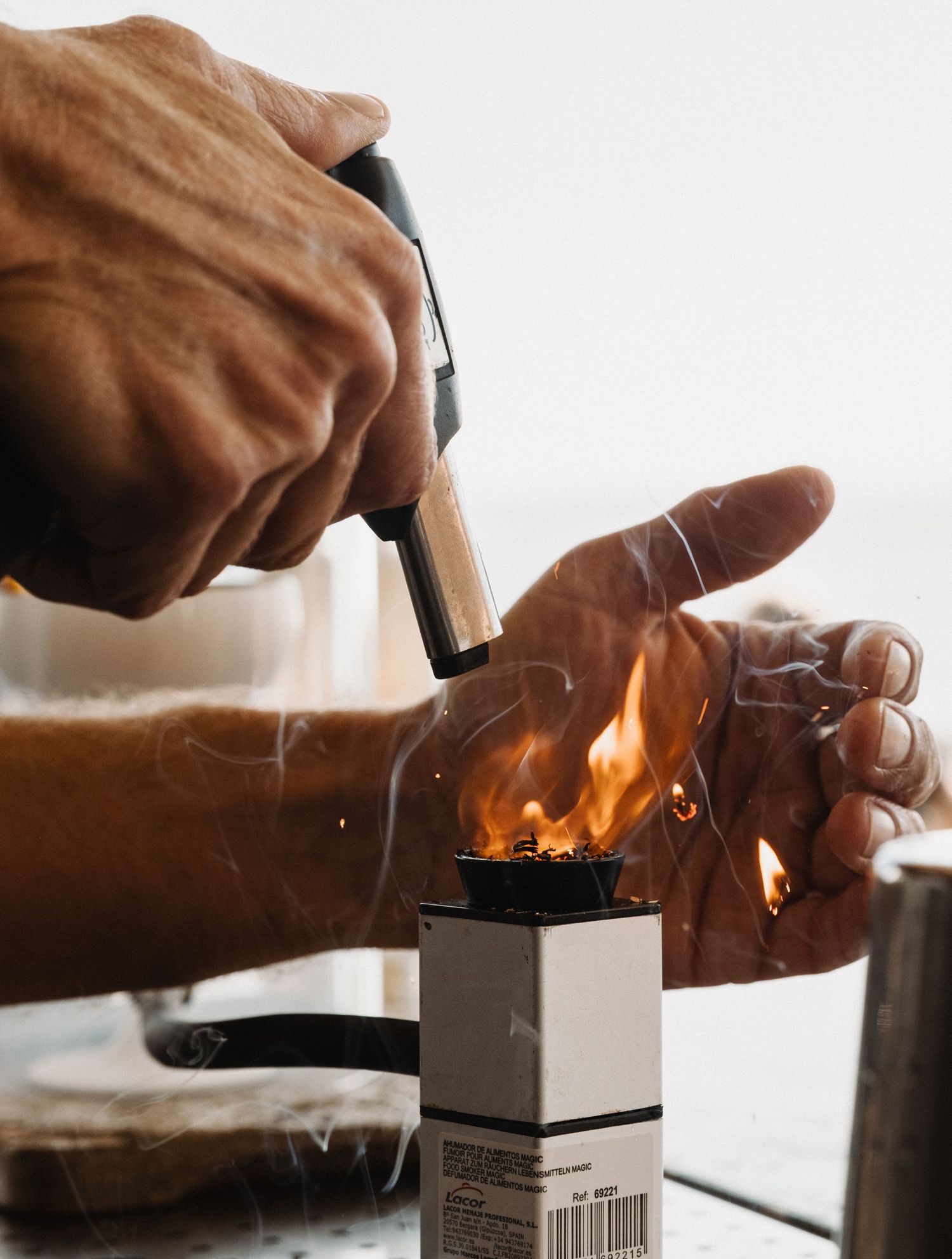 Hands using a blowtorch to ignite flames on a small white box on a table.