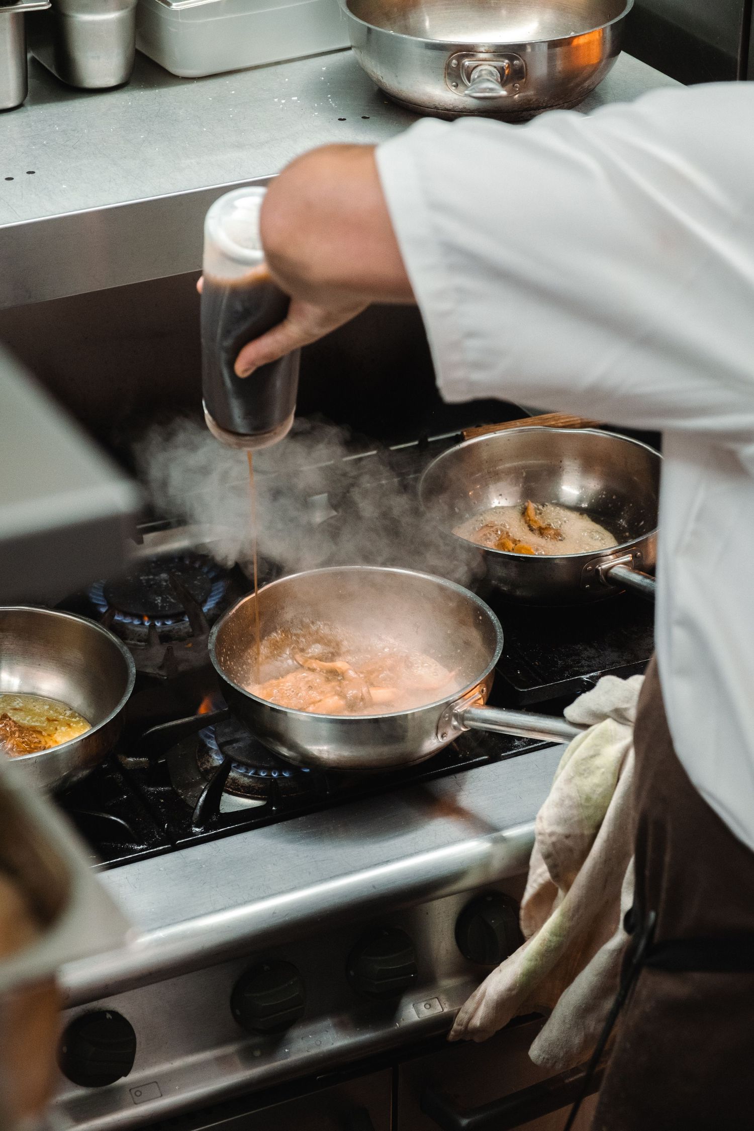Chef cooking in professional kitchen with steaming pans on gas stove.