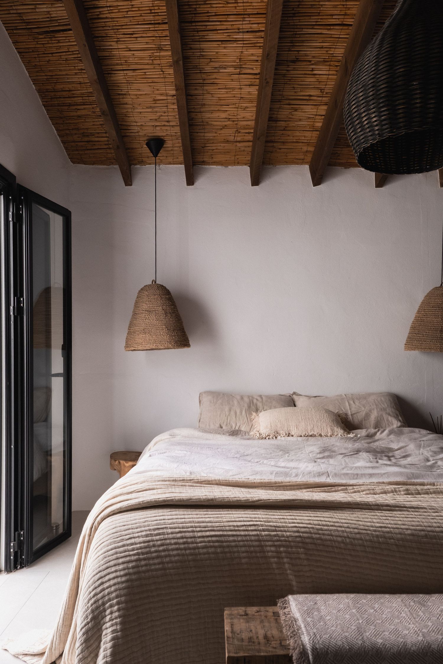 Rustic bedroom with woven pendant lights, exposed wood beam ceiling, neutral linen bedding, and white plaster walls.
