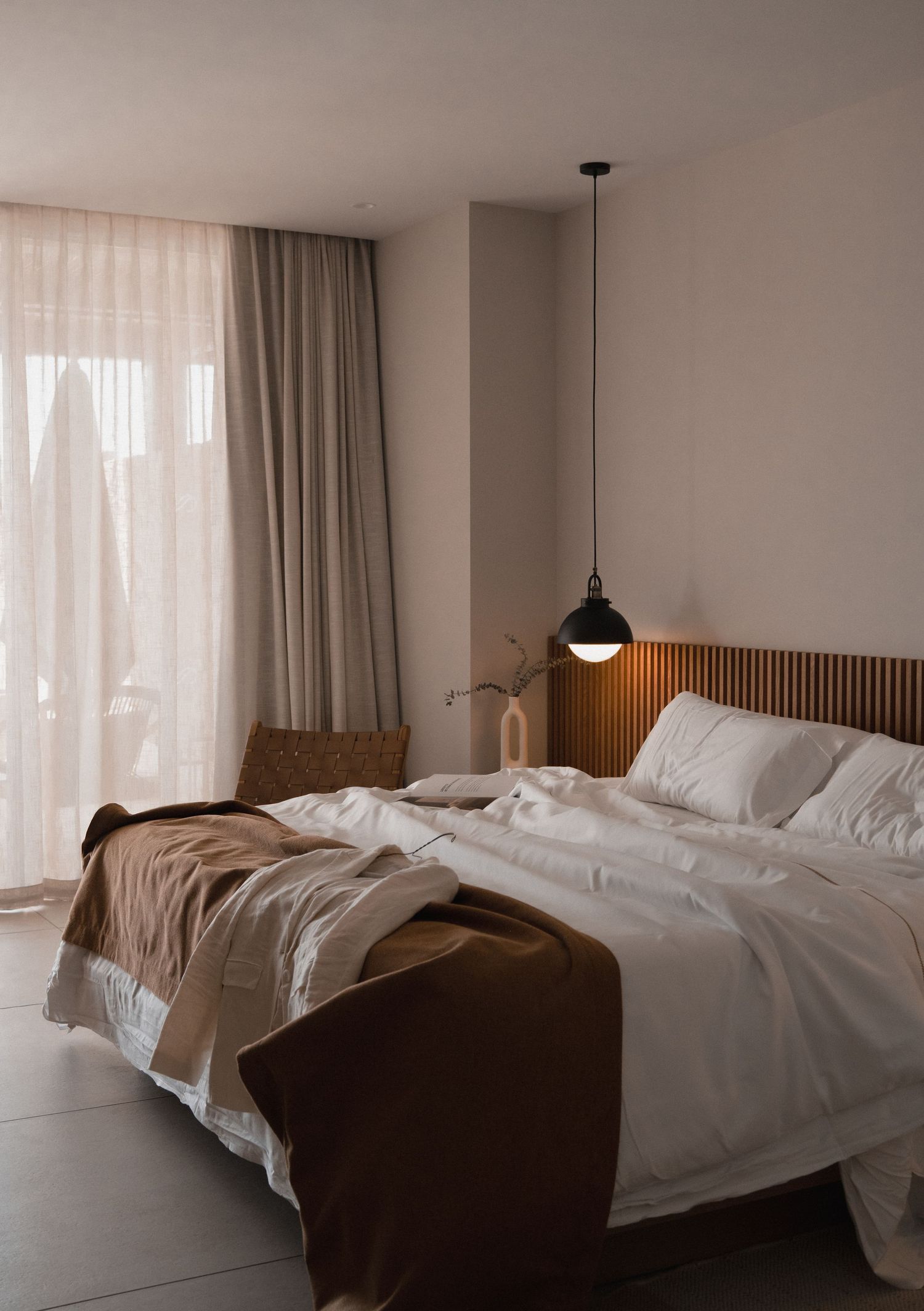 Minimalist bedroom with white bedding, brown accents, pendant light, slatted headboard, and sheer curtains by a large window.