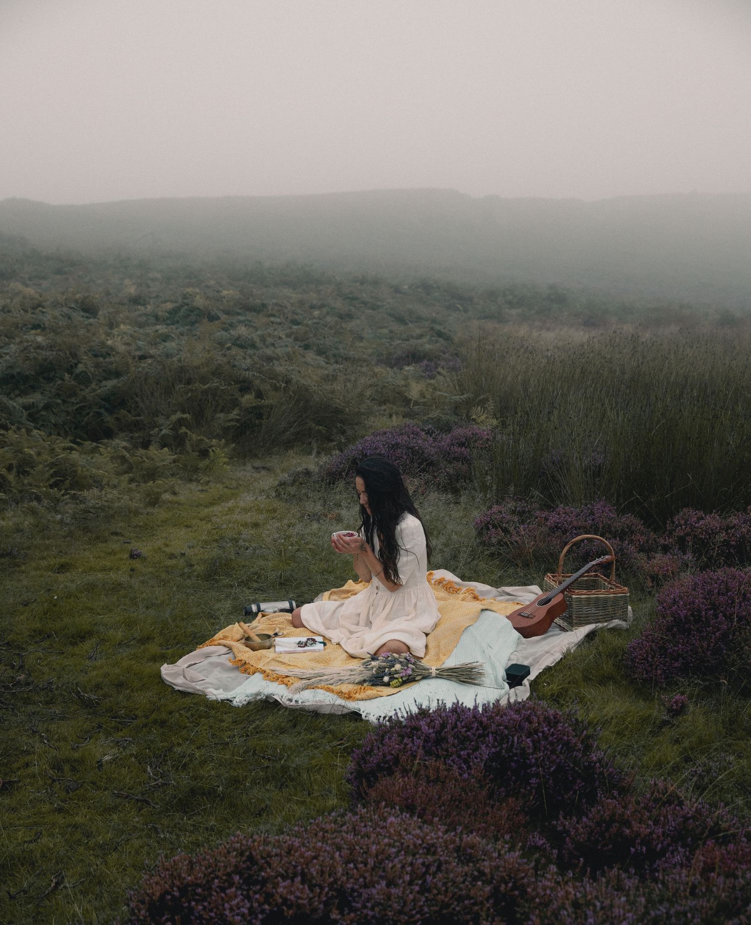 A picnic blanket with food and drinks laid out on misty moorland with purple heather.