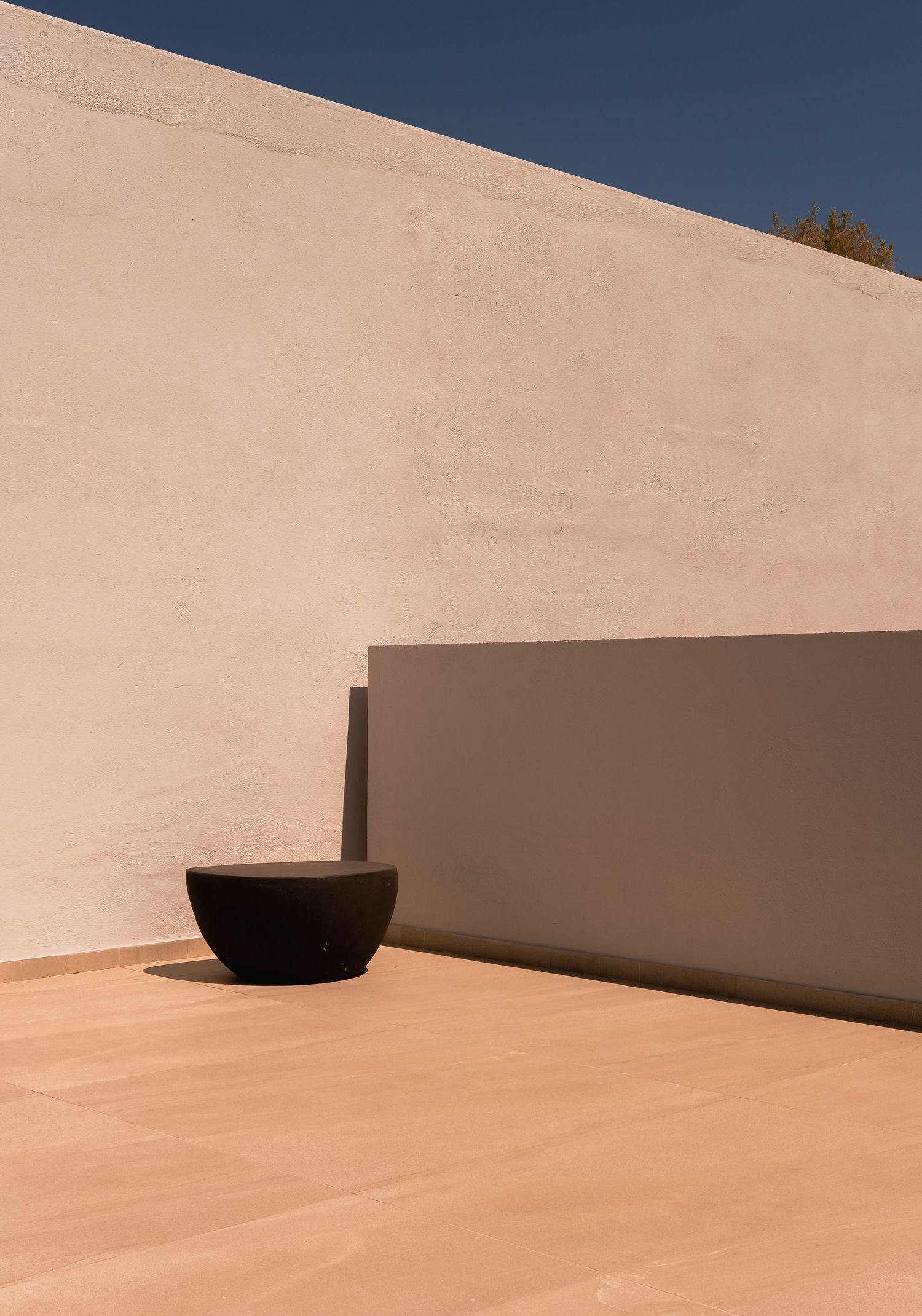Minimalist outdoor courtyard with a black bowl planter against a warm beige stucco wall under a clear blue sky.