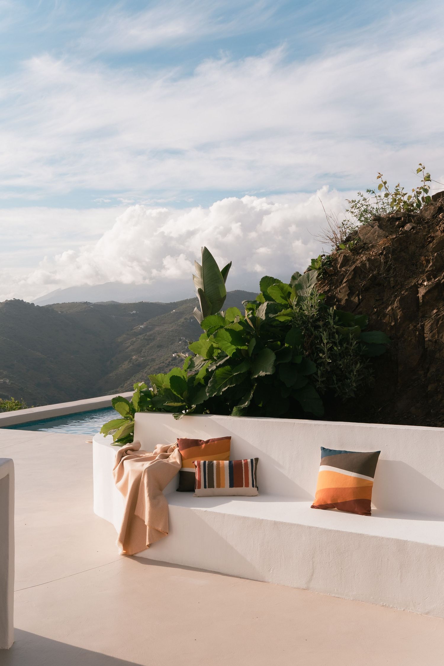 White outdoor bench with colorful pillows and throw overlooking lush mountain valley and infinity pool under cloudy sky.
