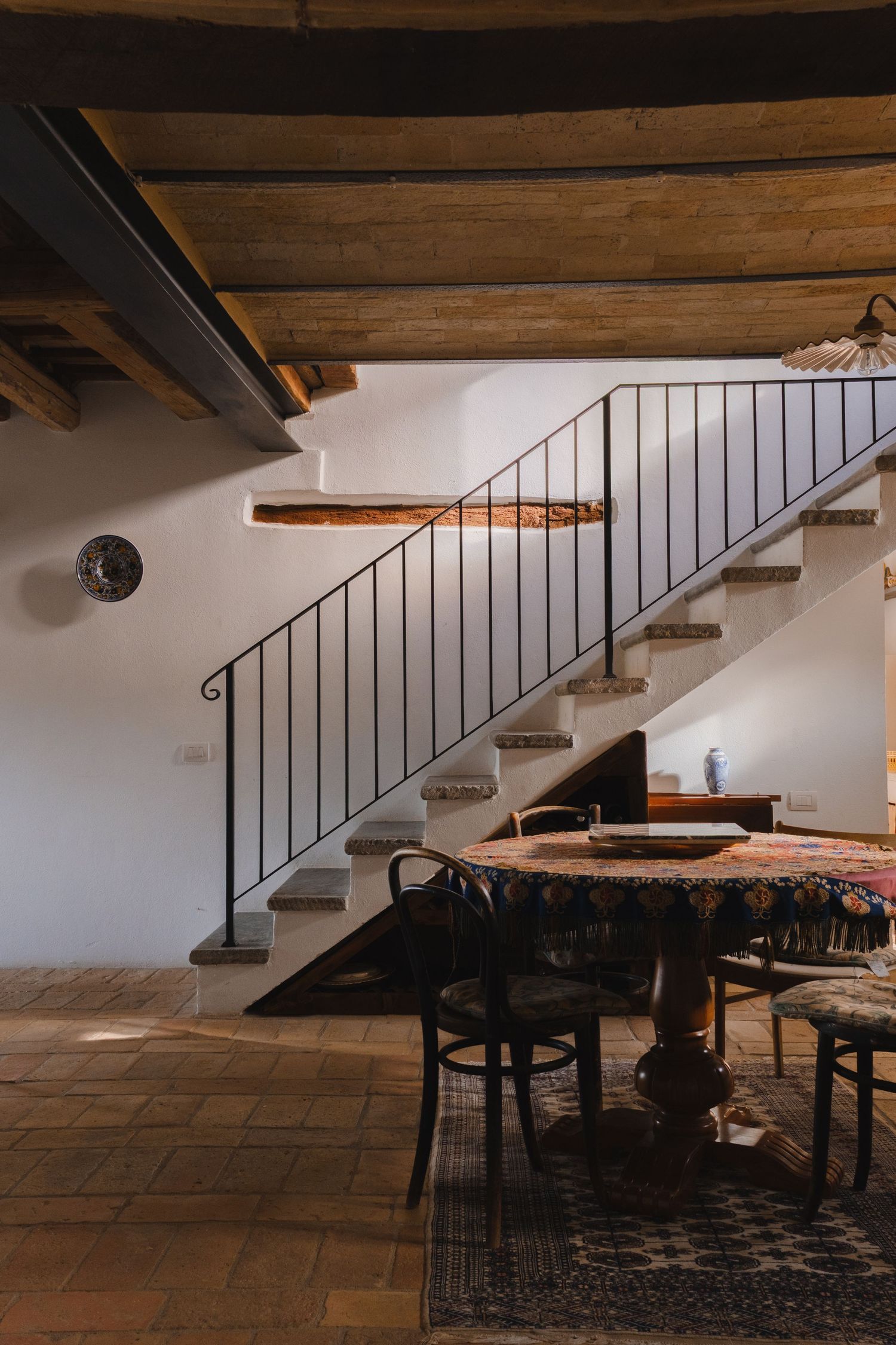 Rustic interior with stone staircase, iron railing, wooden beam ceiling, and antique dining table on terracotta tile floor.