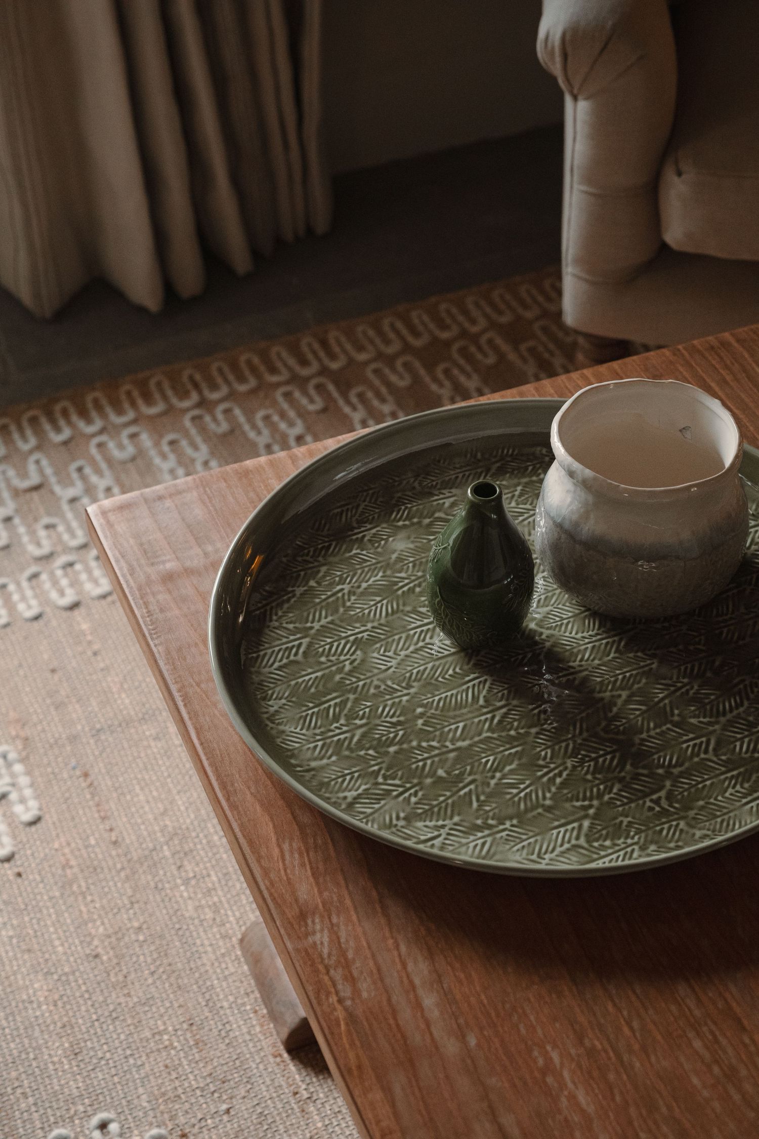 Green textured serving tray with ceramic vessel on wooden table against neutral backdrop.