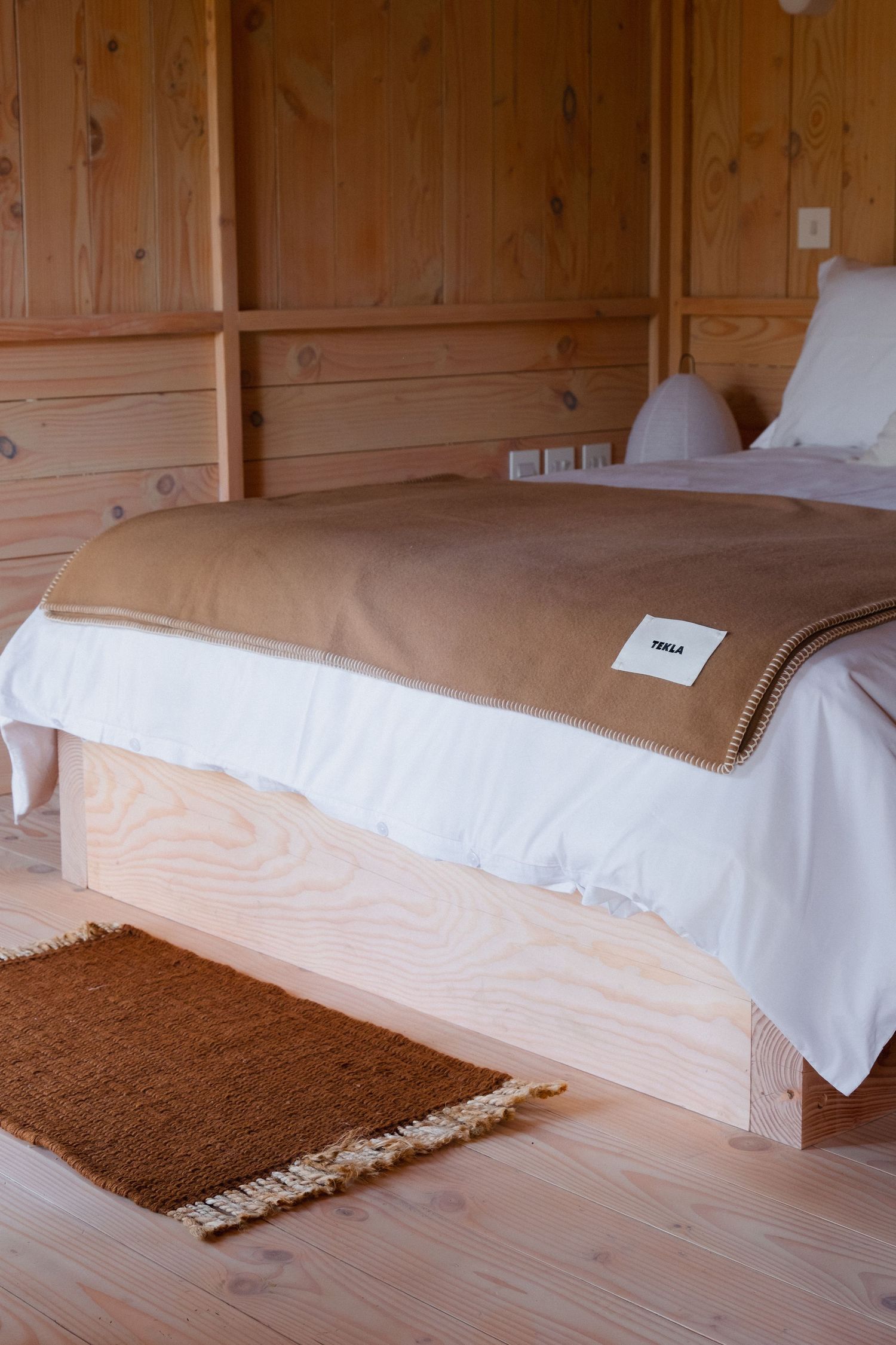 Cozy wooden cabin bedroom with a Tekla blanket on a platform bed, white bedding, and a rust-colored rug.