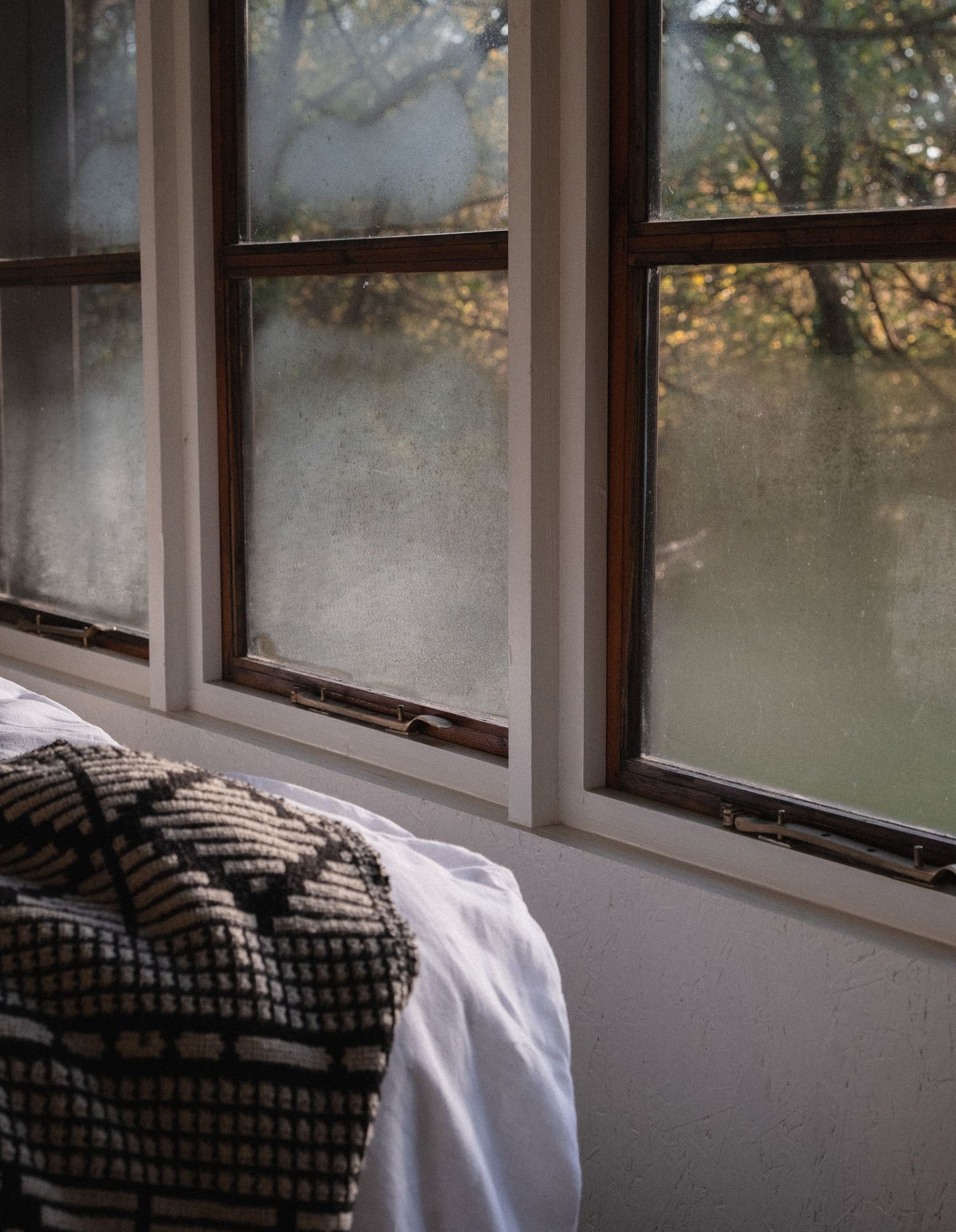 Cozy bedroom corner with patterned blanket near misty wood-framed windows overlooking autumn trees.