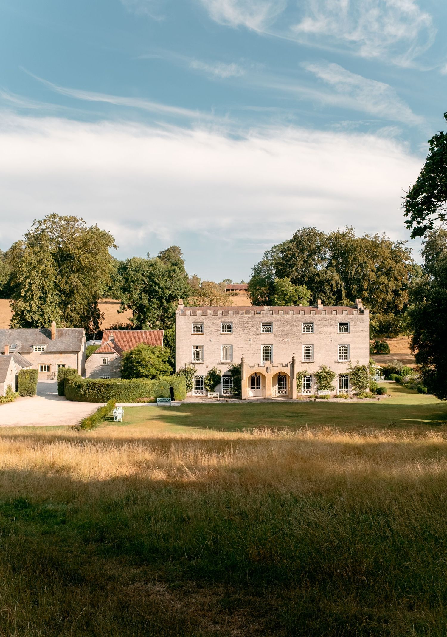 Historic English country manor house surrounded by lush green lawns and mature trees under a blue sky.