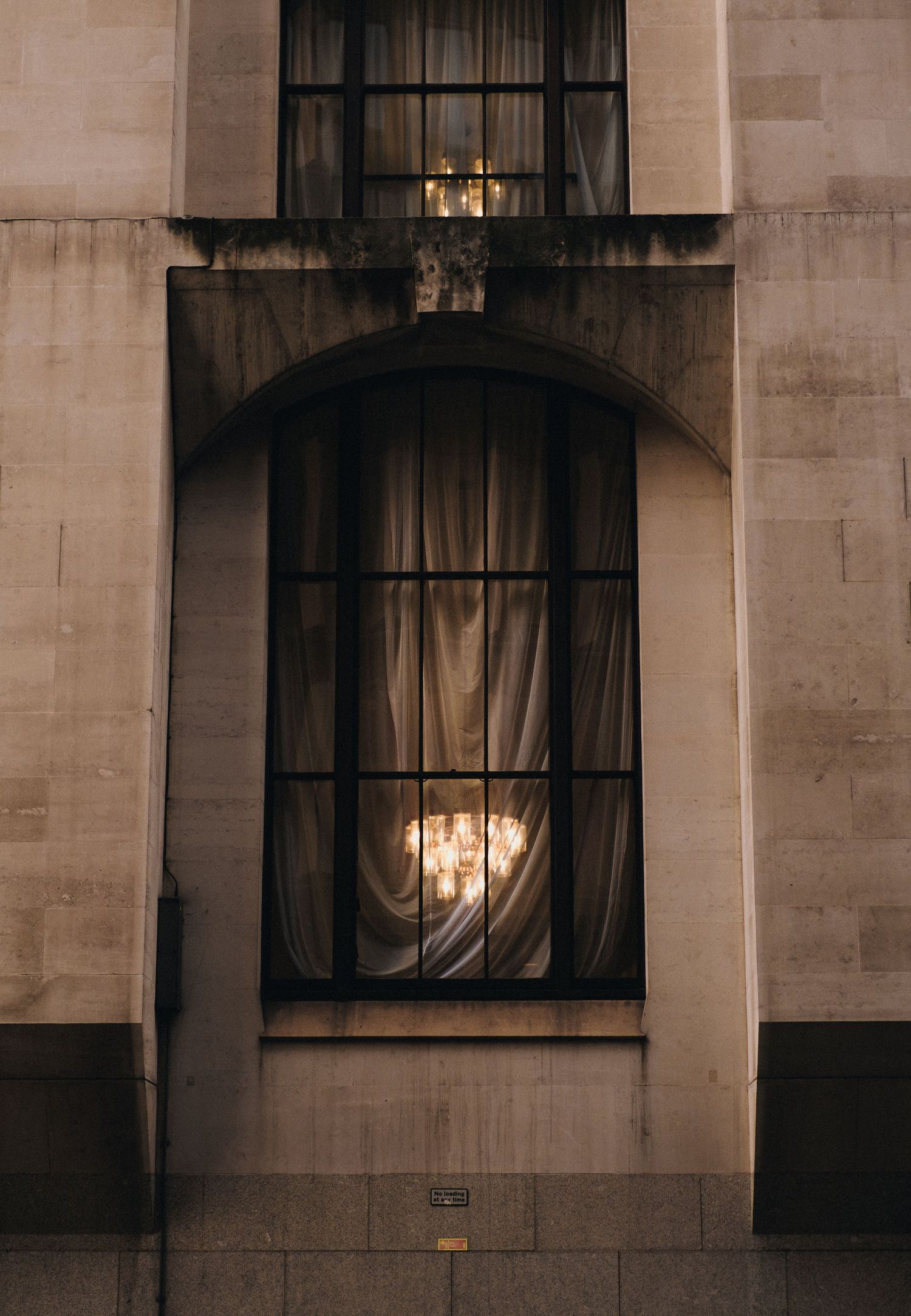 Stone building facade with dark curtained windows and warm interior lighting at dusk.