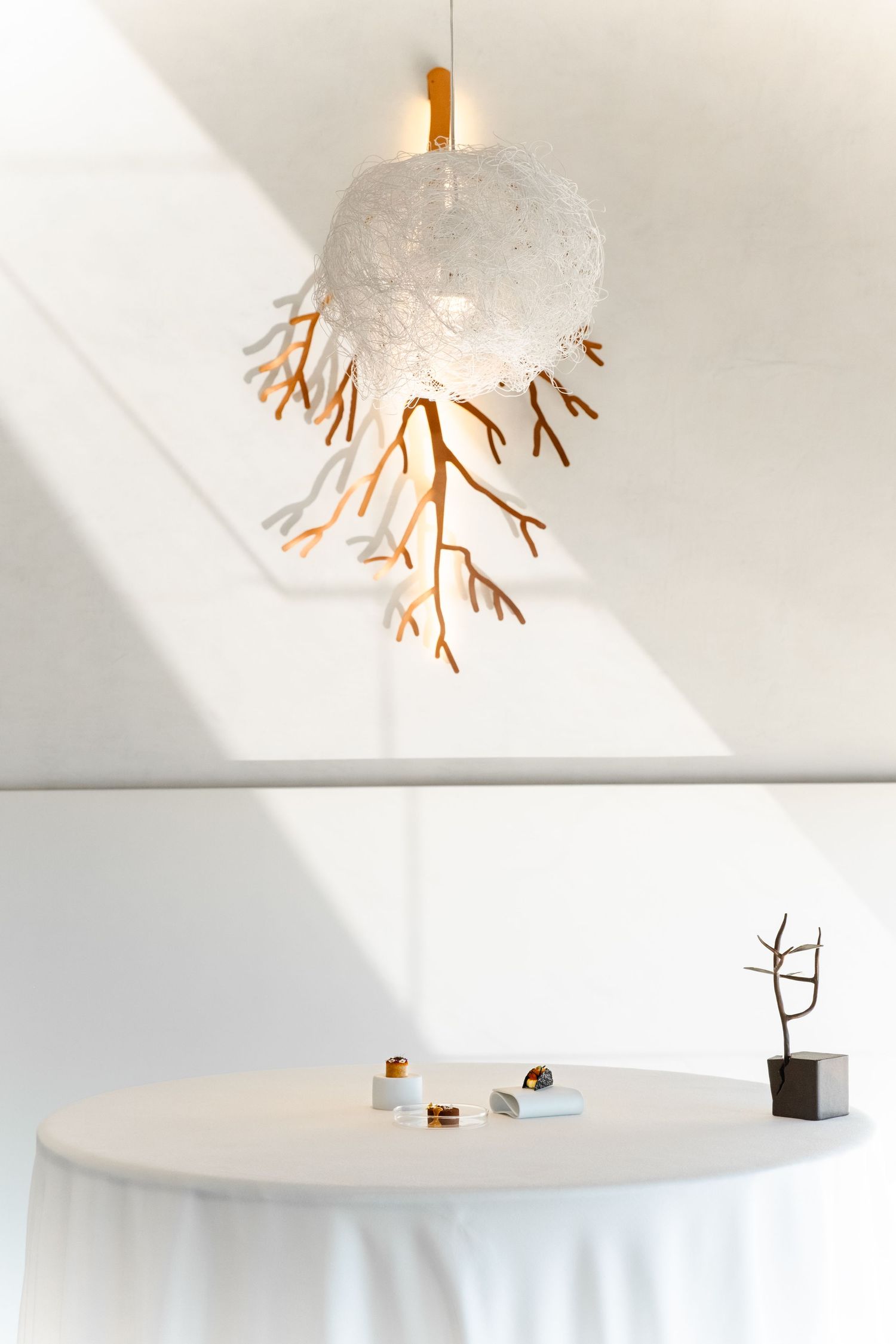Elegant coral-branch pendant light hanging above a white round table with small decorative objects in a bright minimalist space.