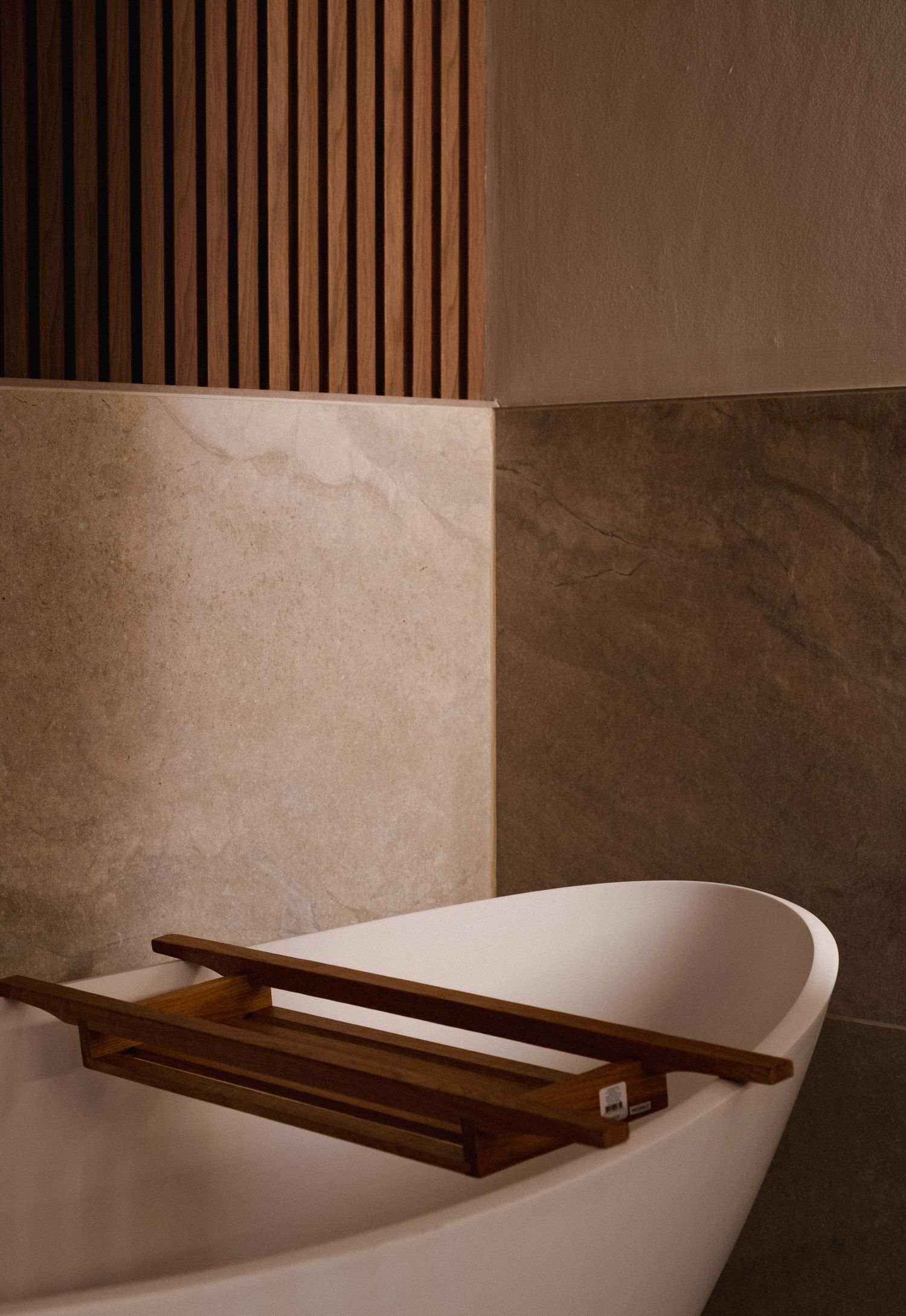 Modern bathroom interior with white freestanding bathtub and wooden tray against brown marble walls and slatted panels.