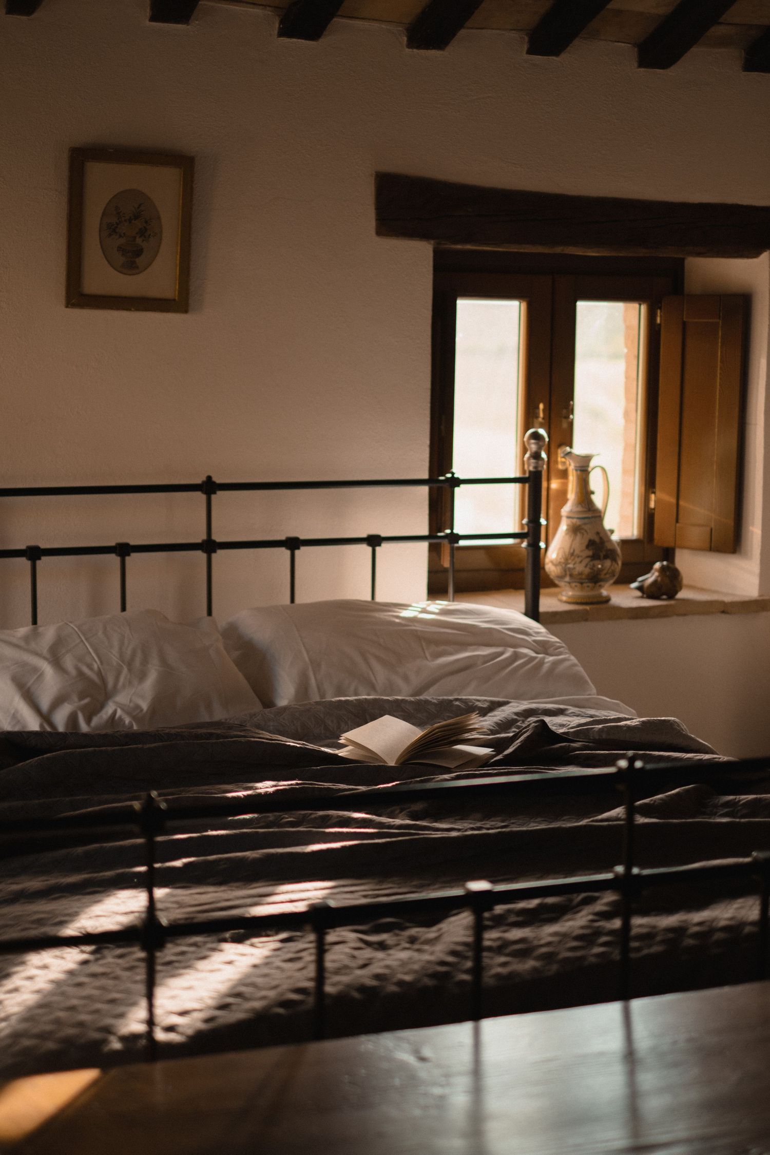 Rustic bedroom with iron bed frame, white pillows, open book, and warm sunlight through wooden window.