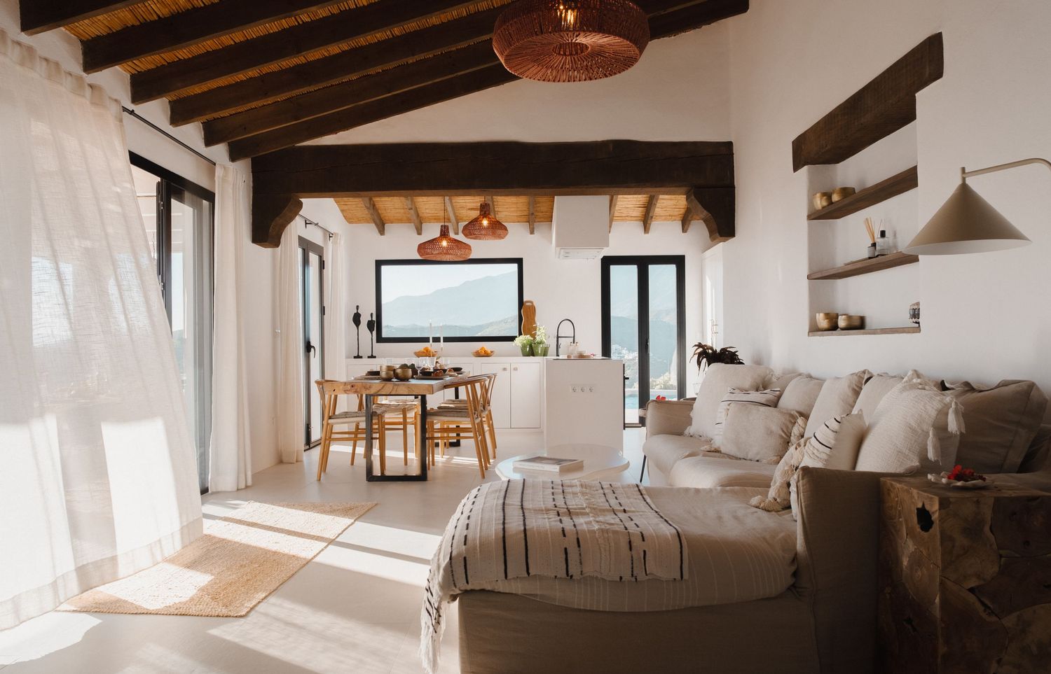 Bright open-plan living room with rustic wooden beams, neutral sofa, and dining area with sea views.
