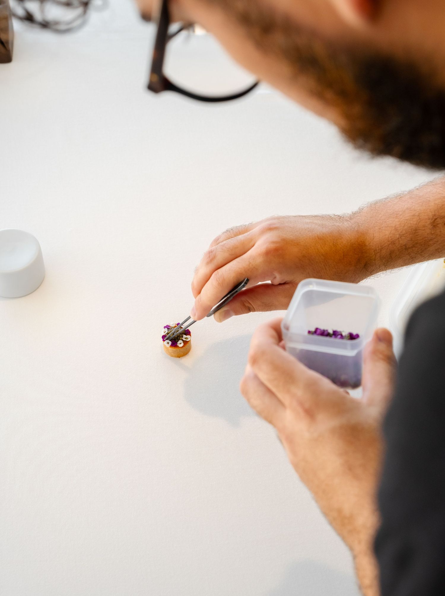 A bearded man uses tweezers to carefully place small purple beads onto a decorative piece at a white workbench.