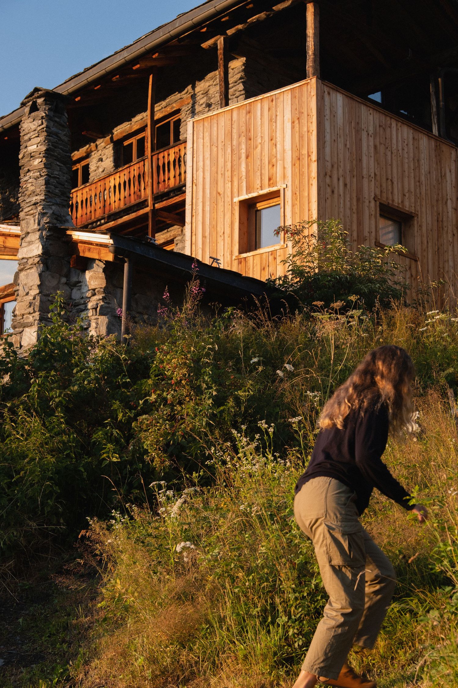 Woman walking through wildflowers beside a rustic wooden alpine chalet bathed in warm golden hour sunlight.