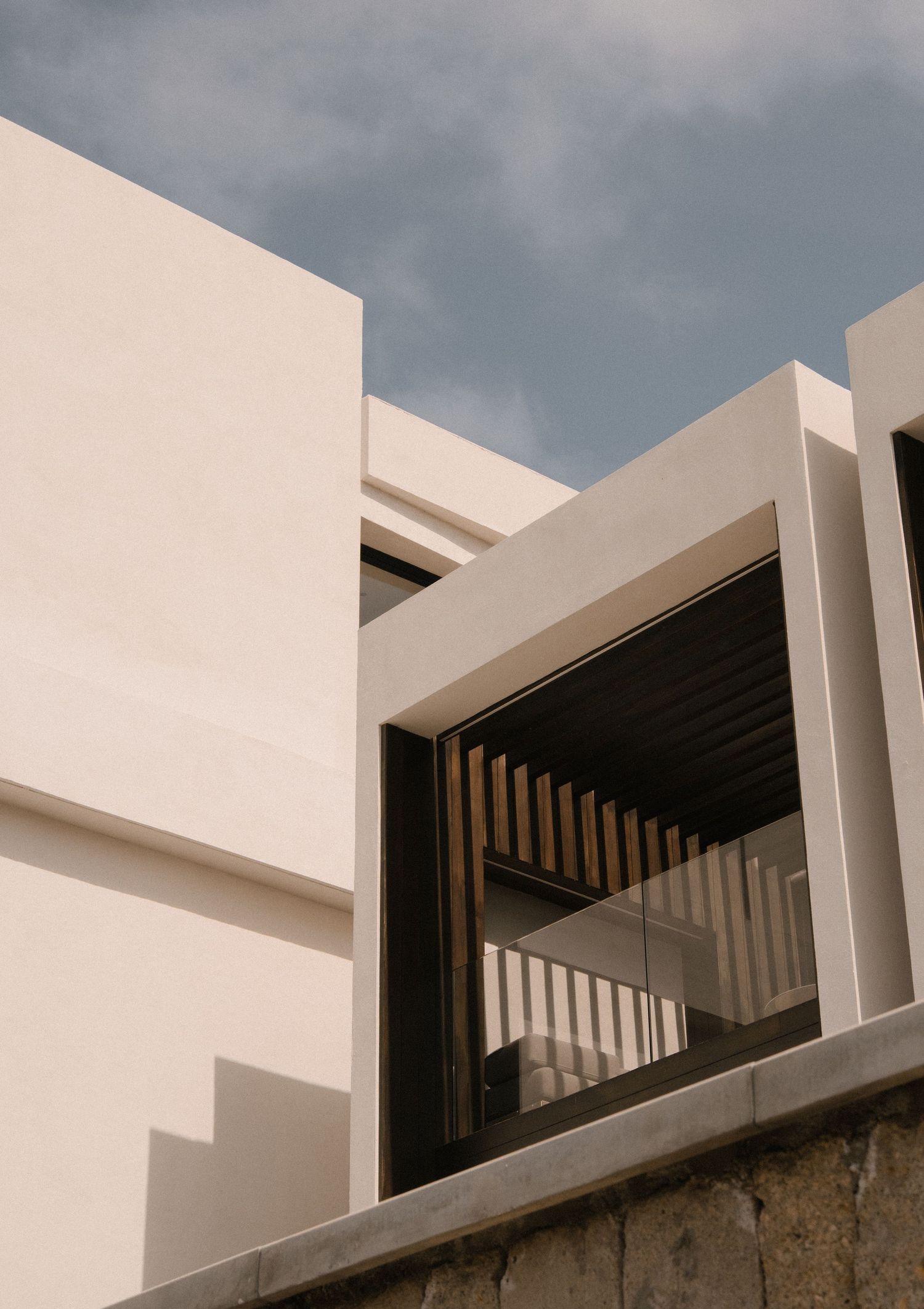 Modern white minimalist building exterior with geometric shapes, wooden louvers, and glass balcony against a blue sky.