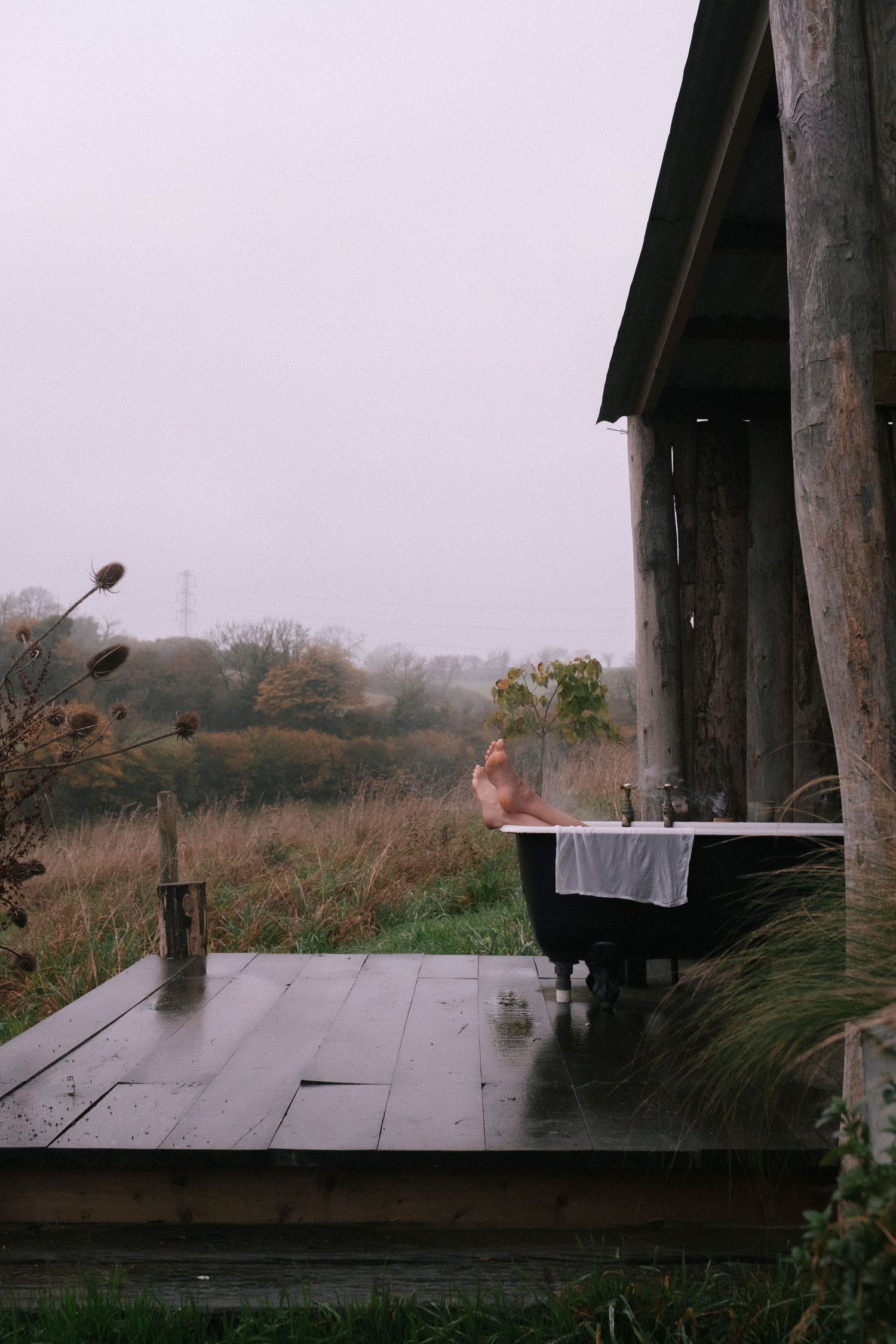 Outdoor clawfoot bathtub on rustic wooden deck overlooking misty rural landscape at dusk.